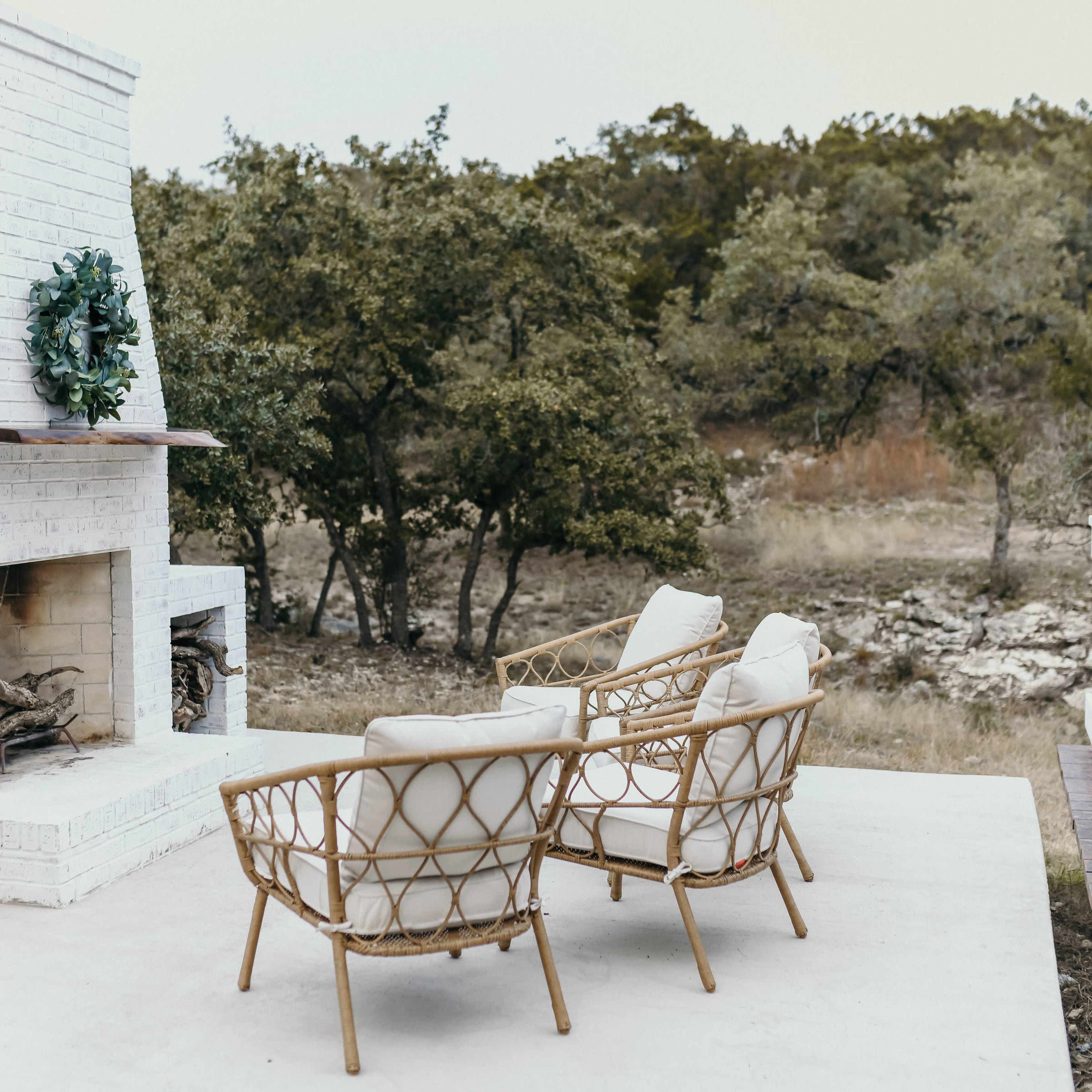 A white outdoor fireplace with a wreath sits between two rattan chairs on a patio overlooking a wooded area.