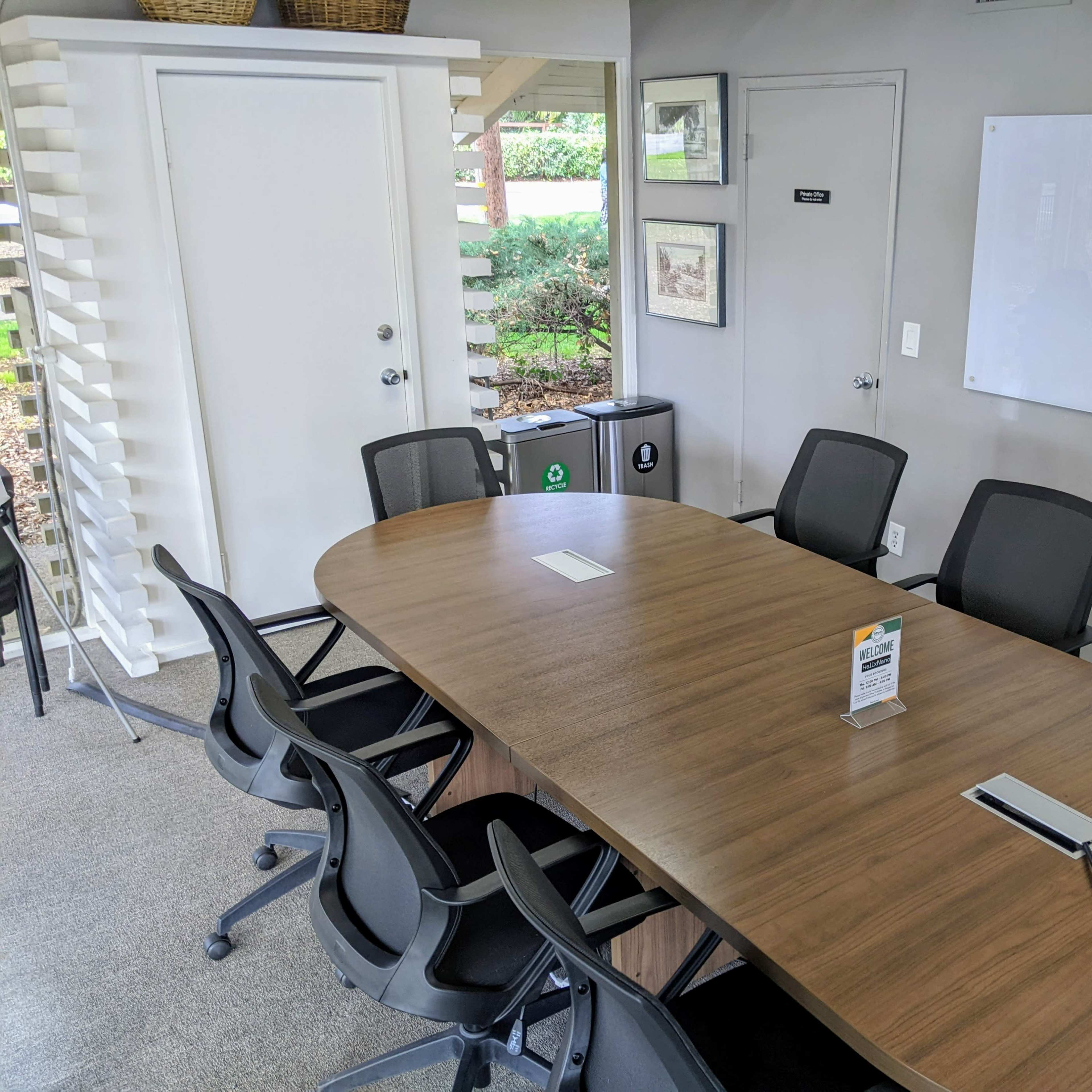 A conference room features a large wooden table surrounded by black office chairs, with a whiteboard and a door leading to a landscaped area visible in the background.