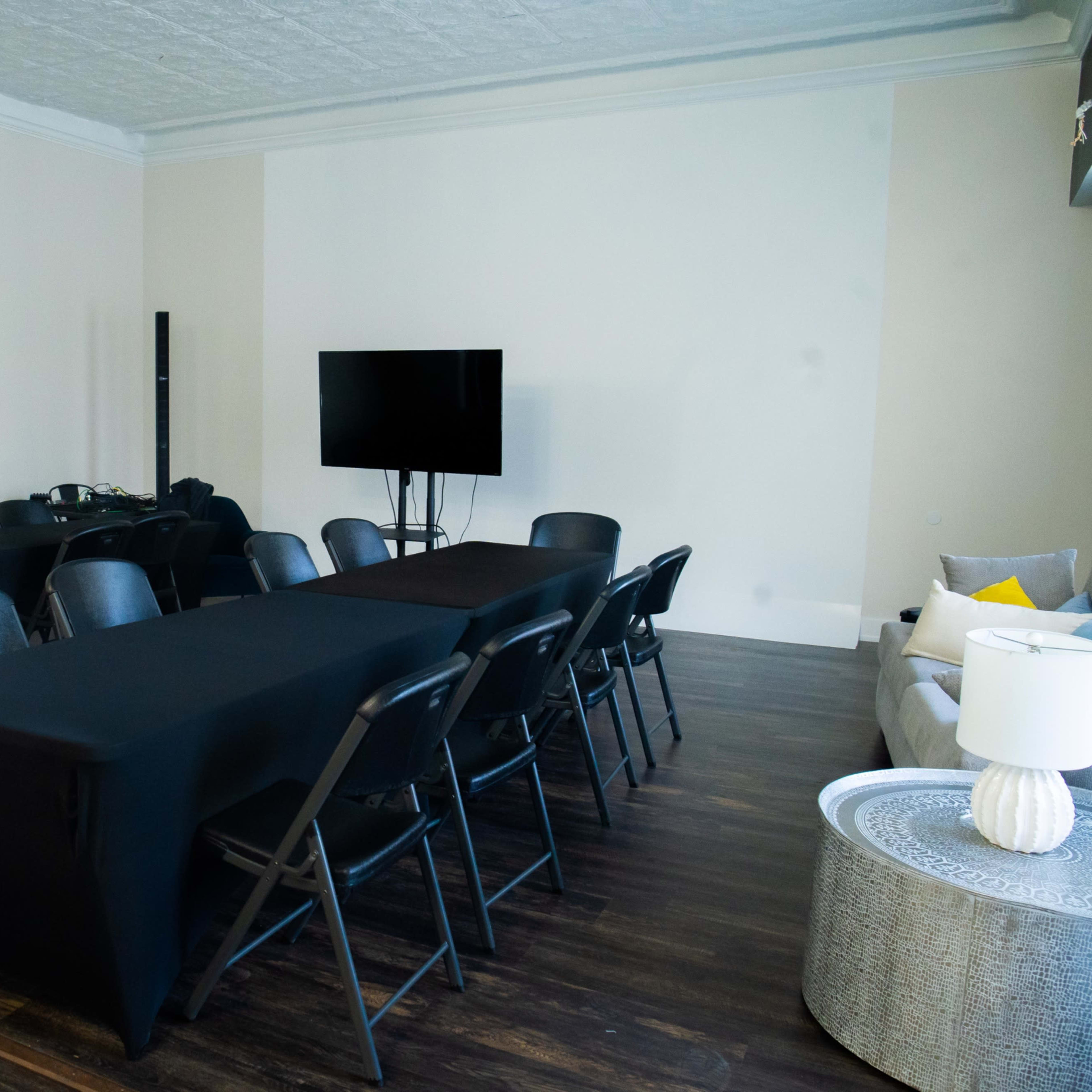 A conference room is set up with several black tables and chairs arranged in a rectangular layout, featuring a television mounted on the wall.