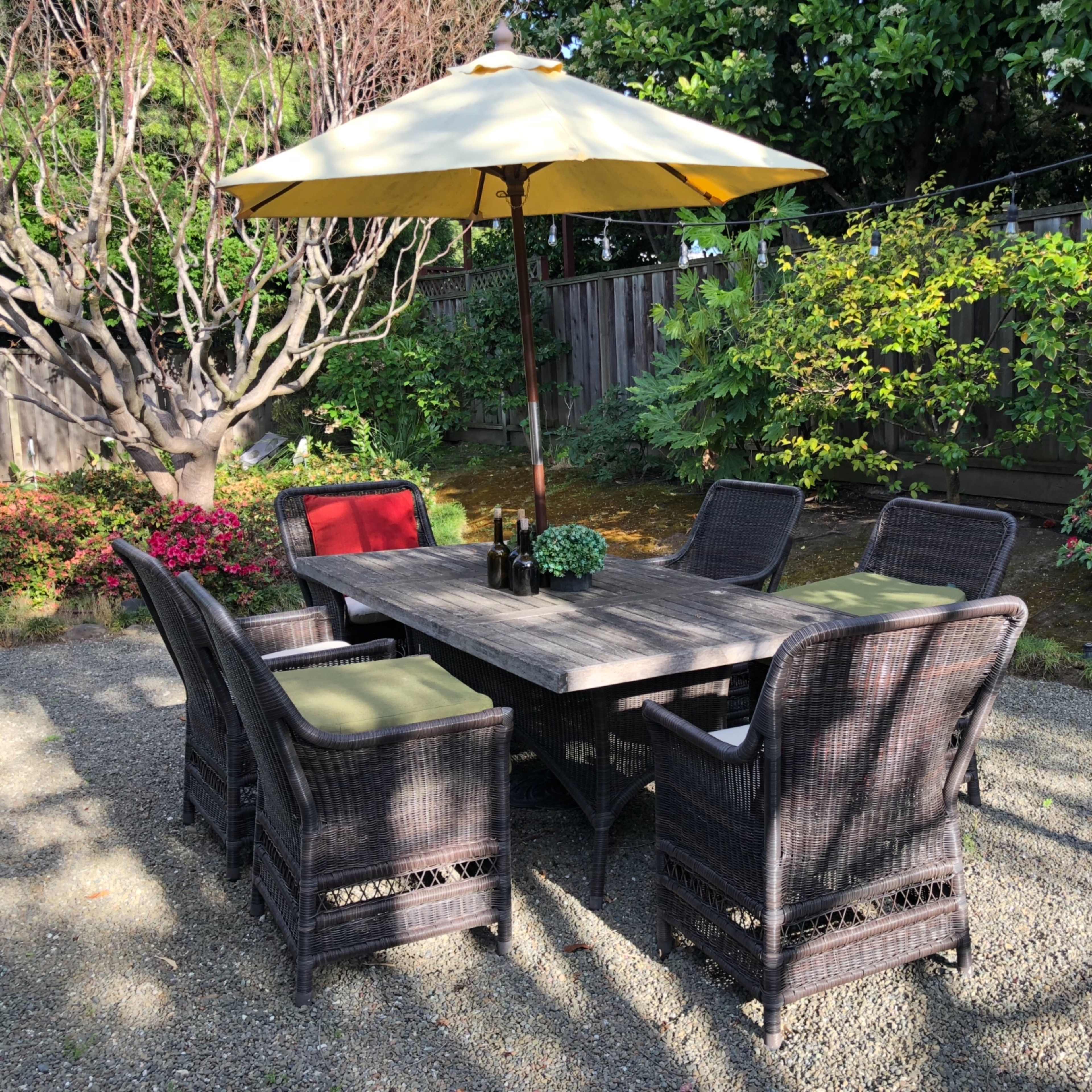 A patio set with a large yellow umbrella stands in a gravel area surrounded by greenery and shrubs.