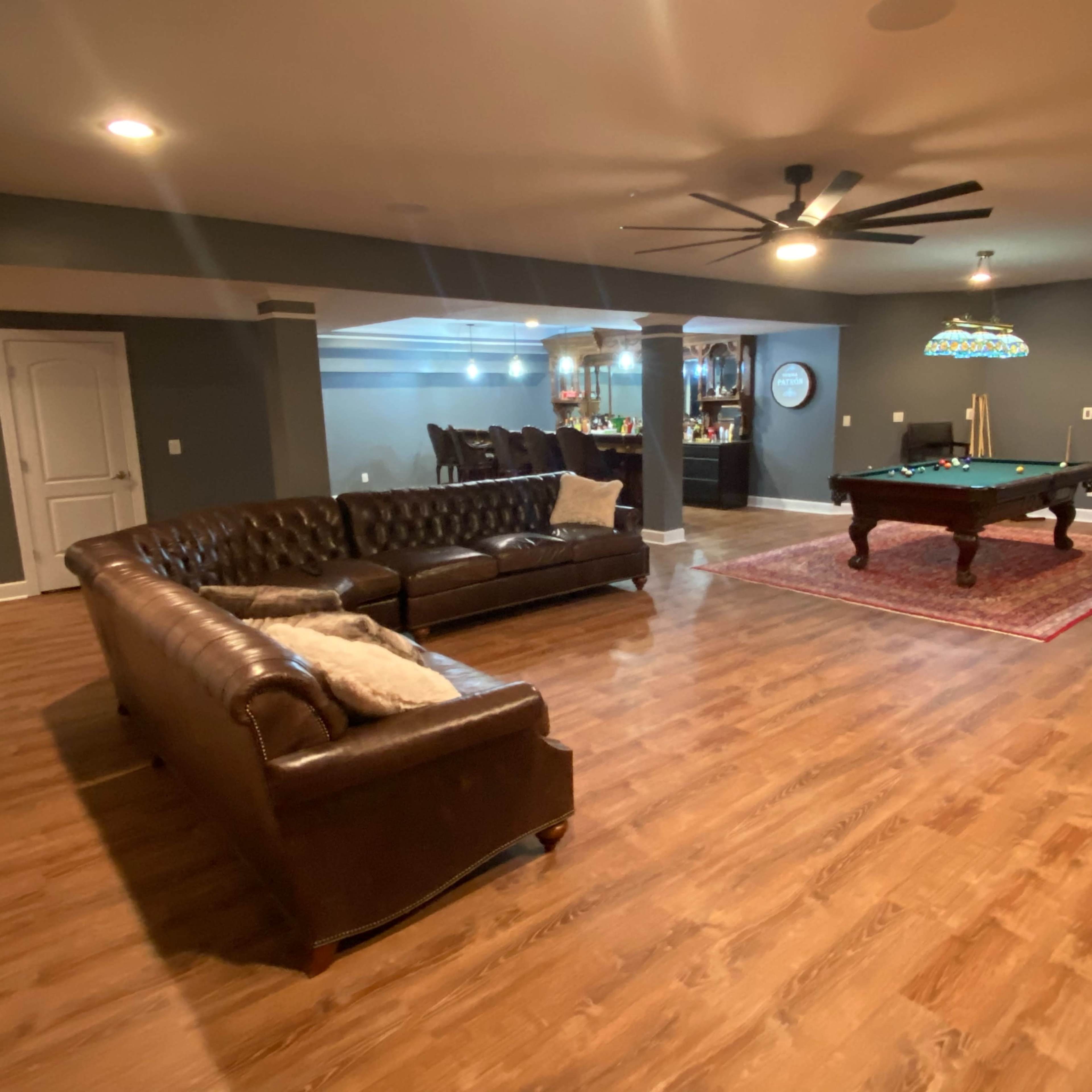 A finished basement with a pool table, a brown leather sectional sofa, and a bar area in the background.