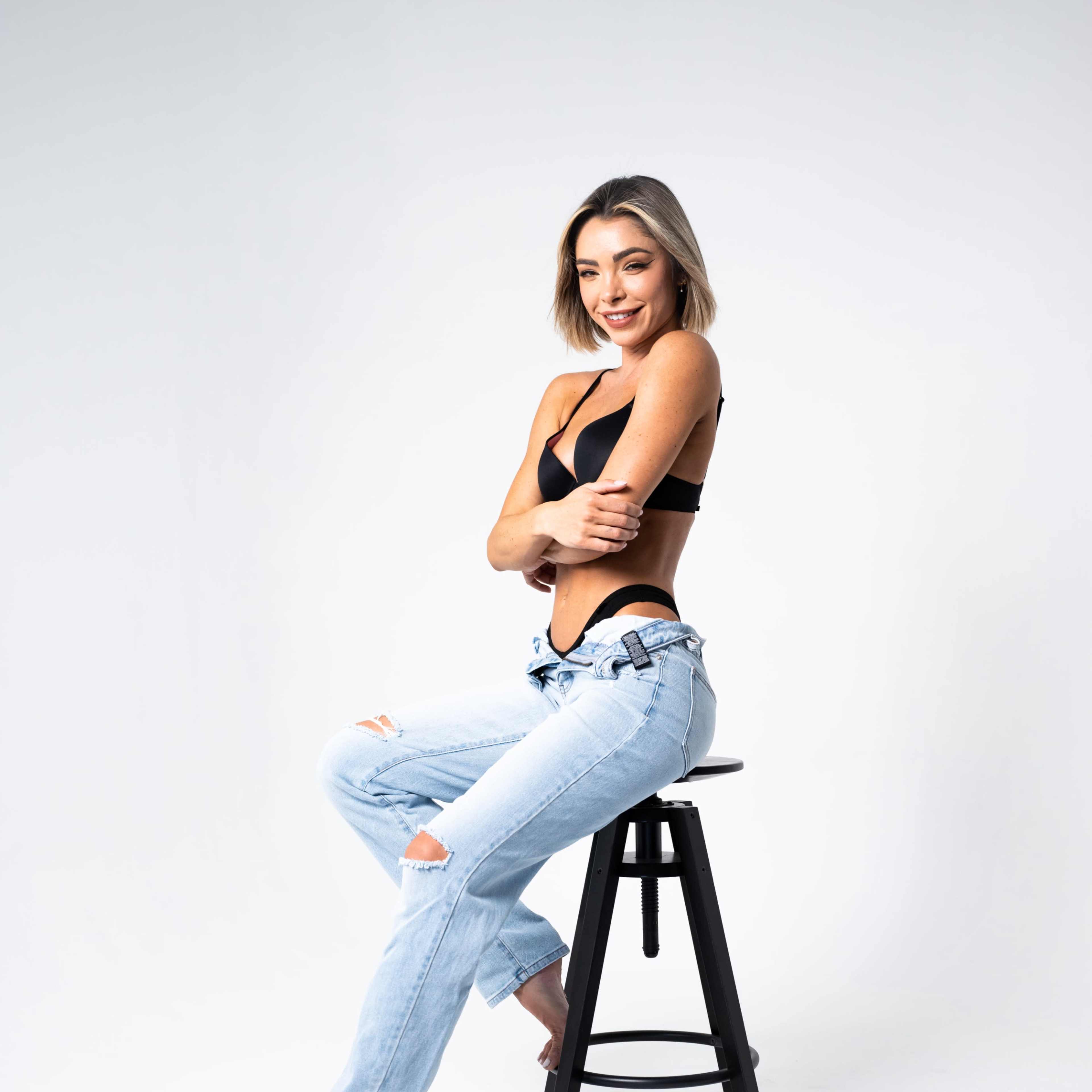 A woman in a black top and ripped light-wash jeans sits on a black stool against a plain background.
