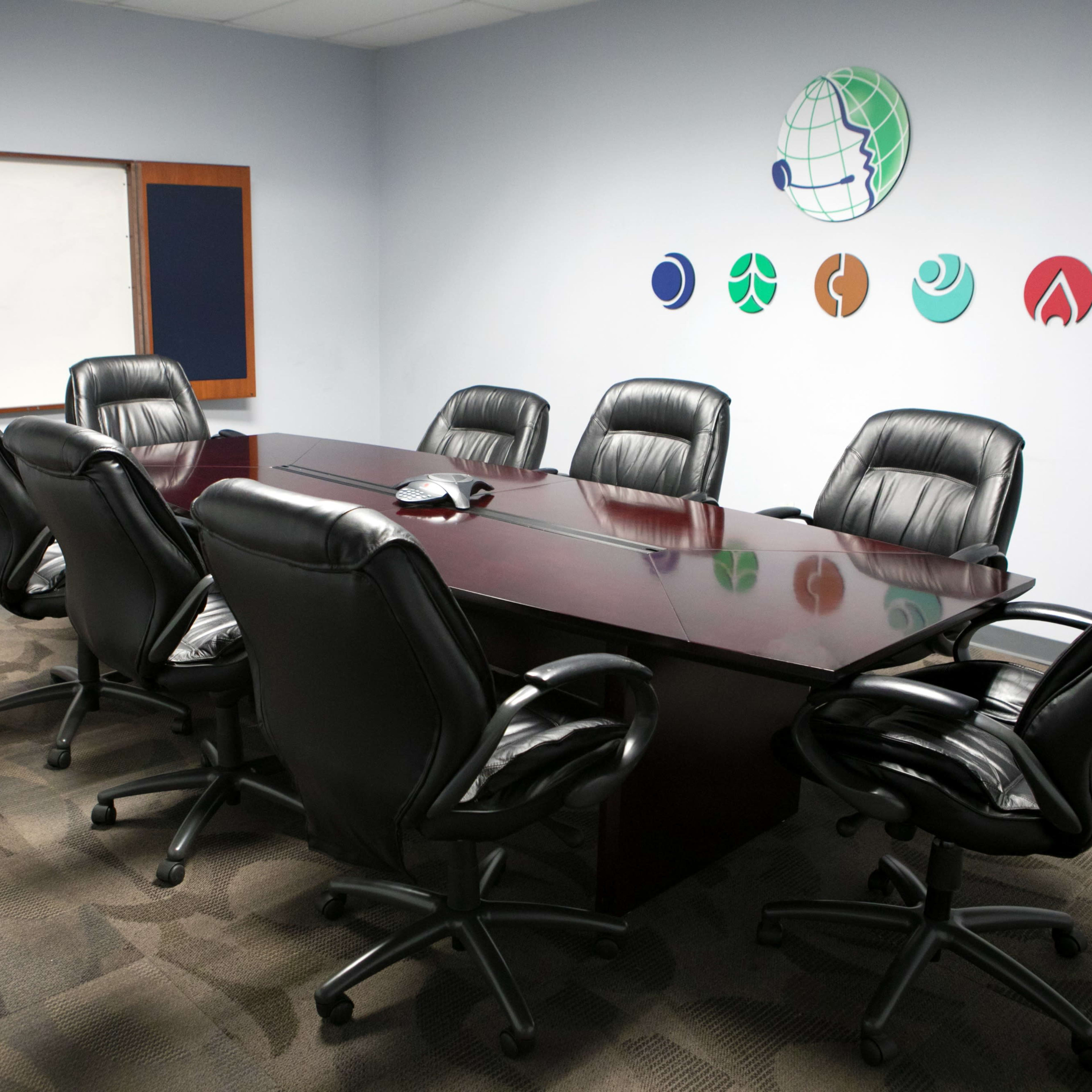A conference room features a long wooden table surrounded by black leather chairs, with wall decorations of various logos.