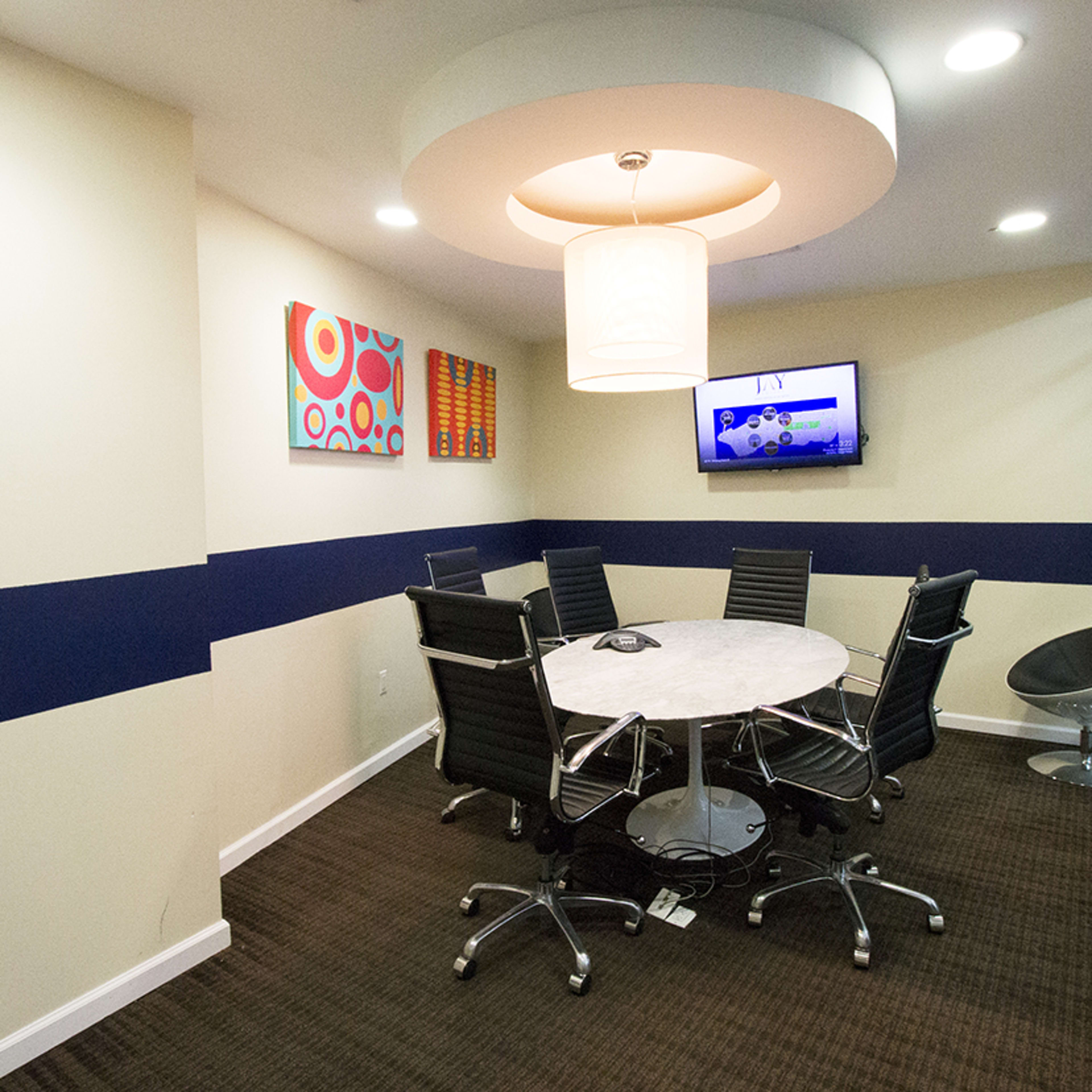A modern meeting room features a round table surrounded by black chairs, a large circular light fixture, and a wall-mounted television.