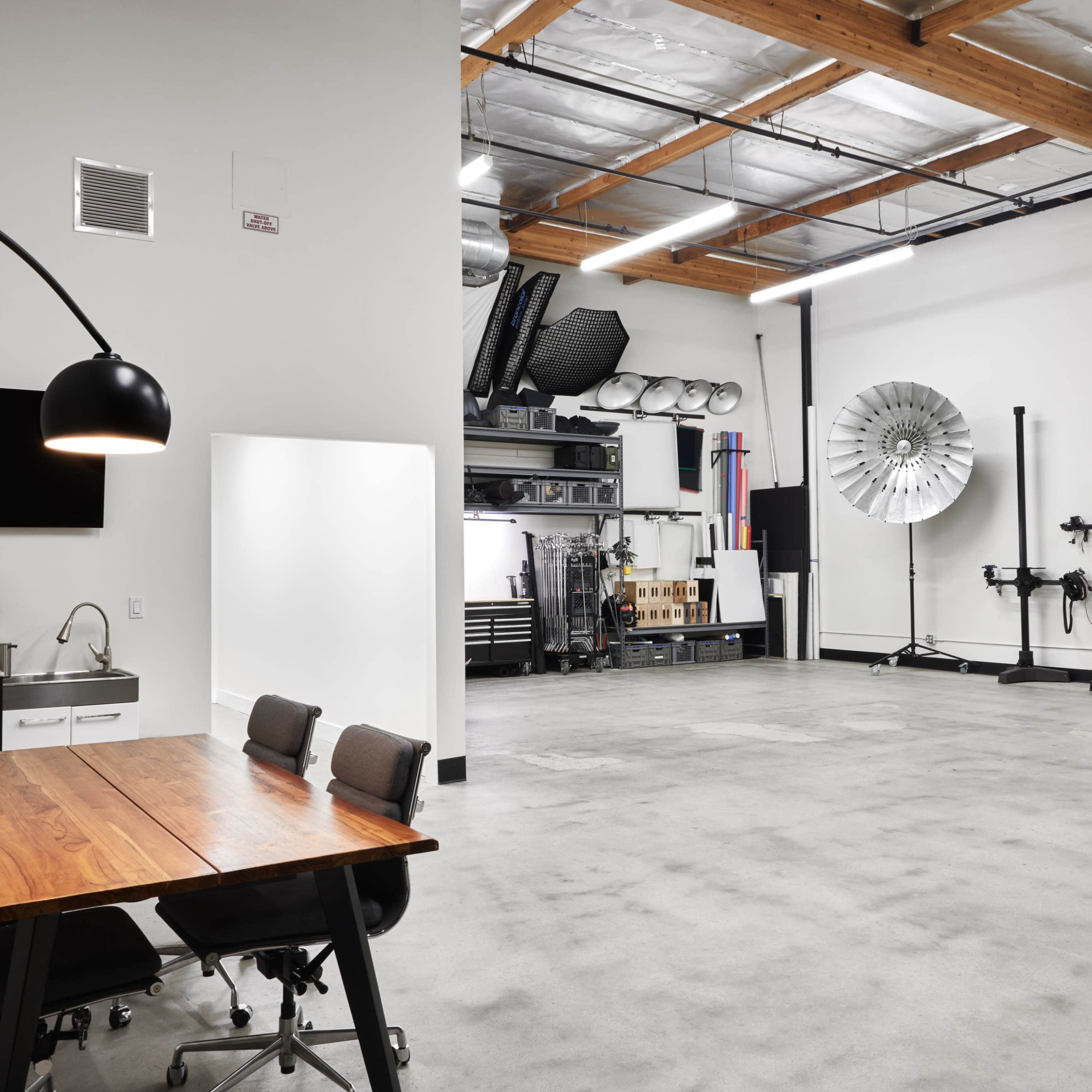 A spacious, minimally furnished studio with a large table and chairs on one side and photography equipment on the other.