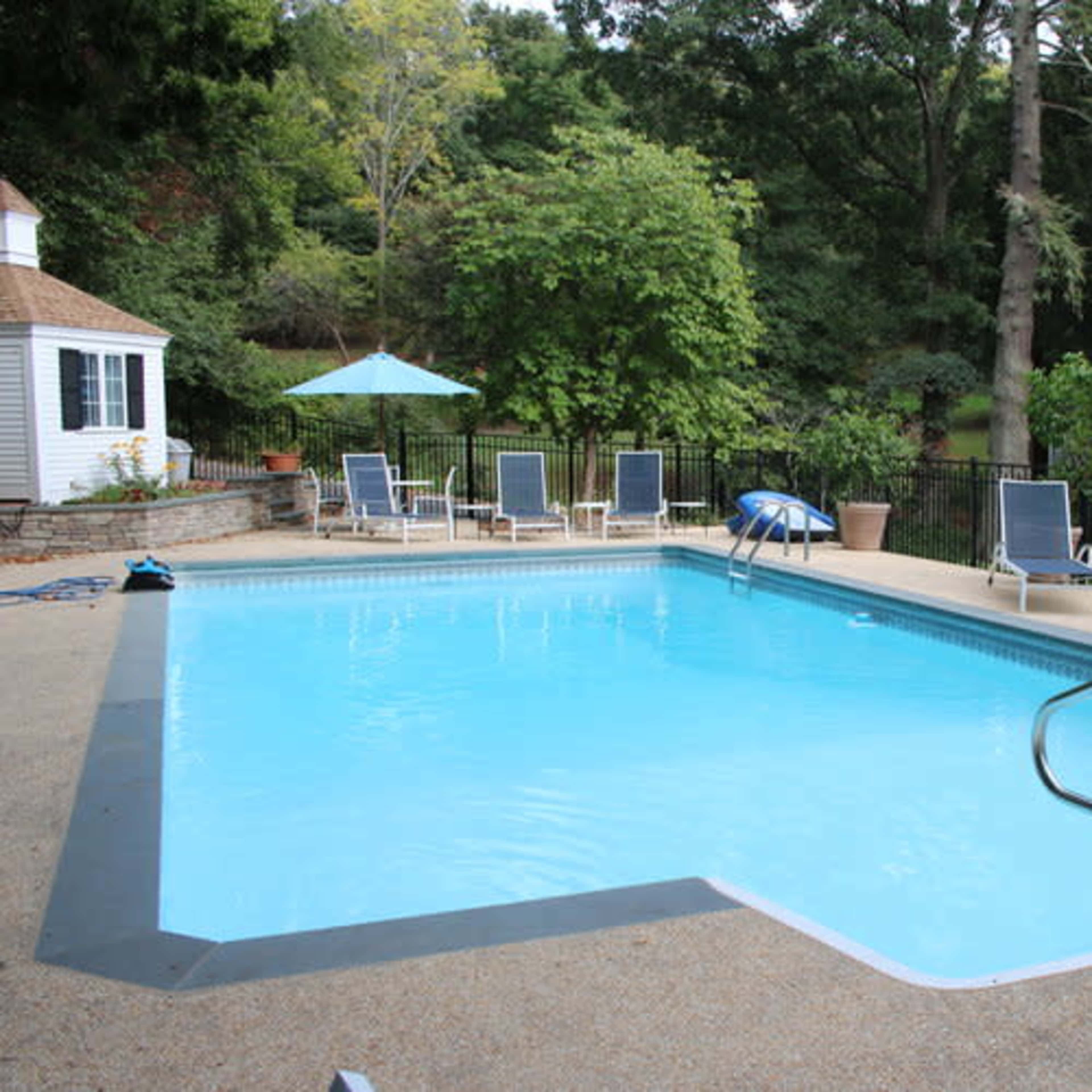A rectangular swimming pool is surrounded by lounge chairs and patio furniture in a lush backyard.