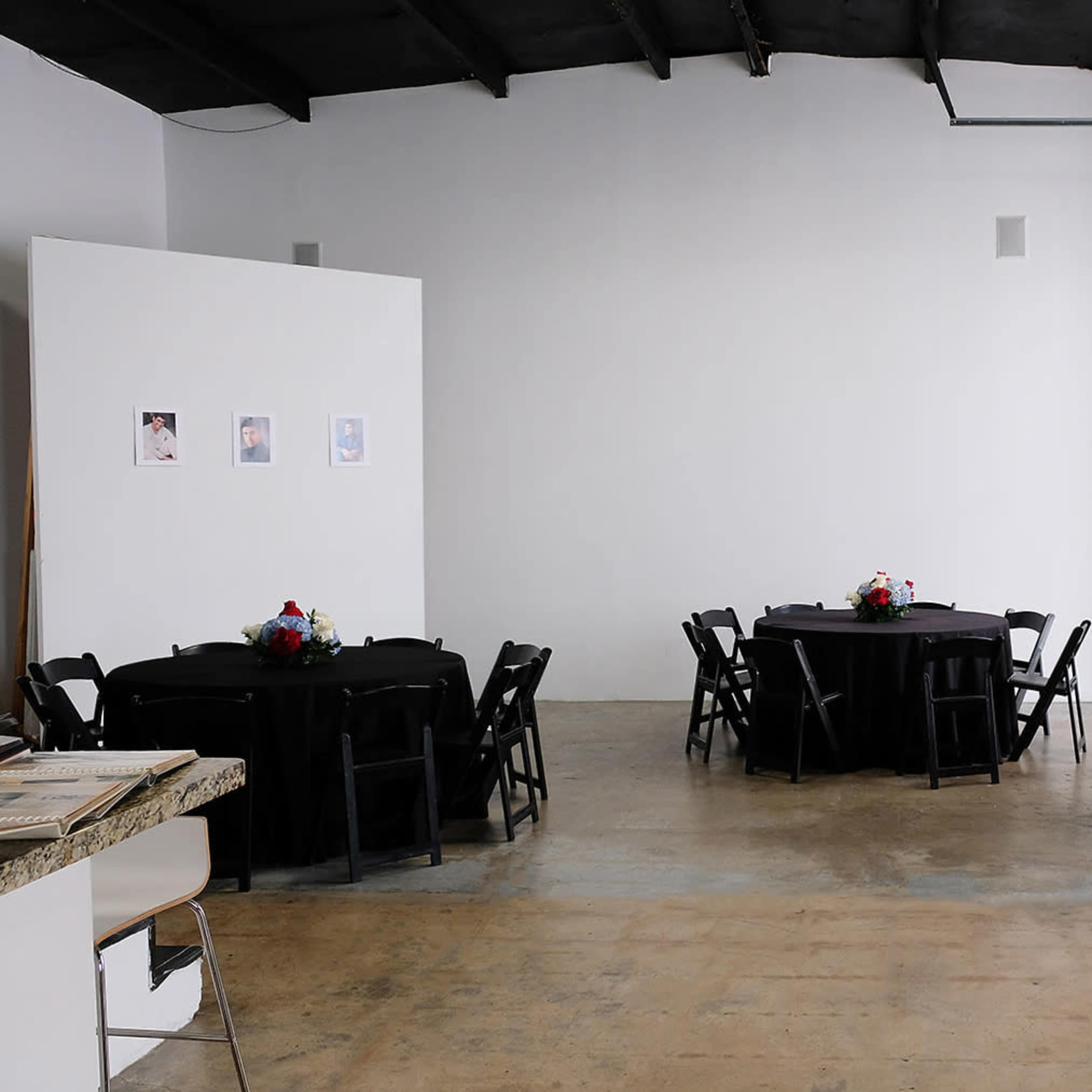 A spacious event venue with two round tables covered in black cloth, each adorned with floral centerpieces, set against a plain white wall.