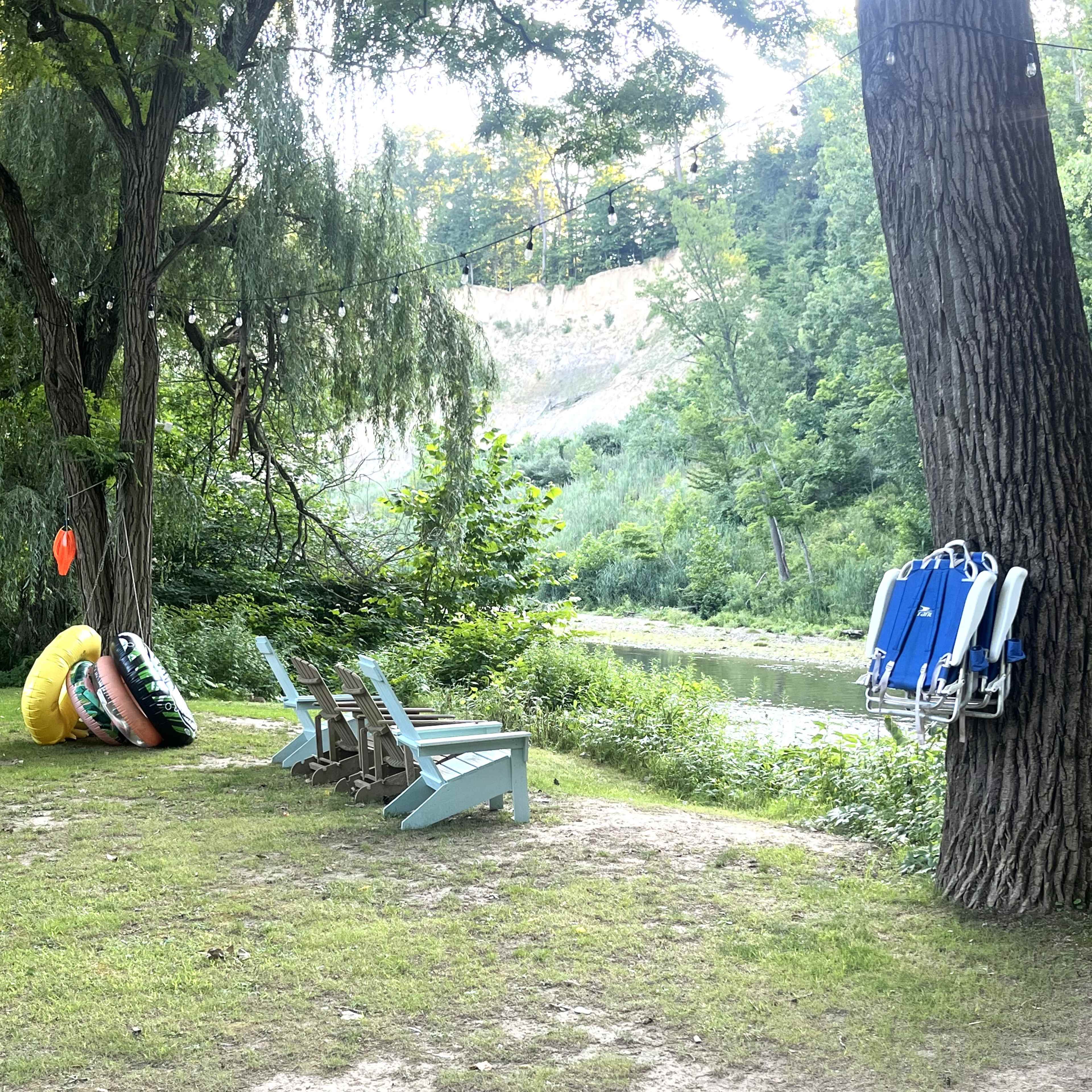 A grassy area beside a river features a set of adirondack chairs and colorful inflatable flotation devices hanging from a tree.