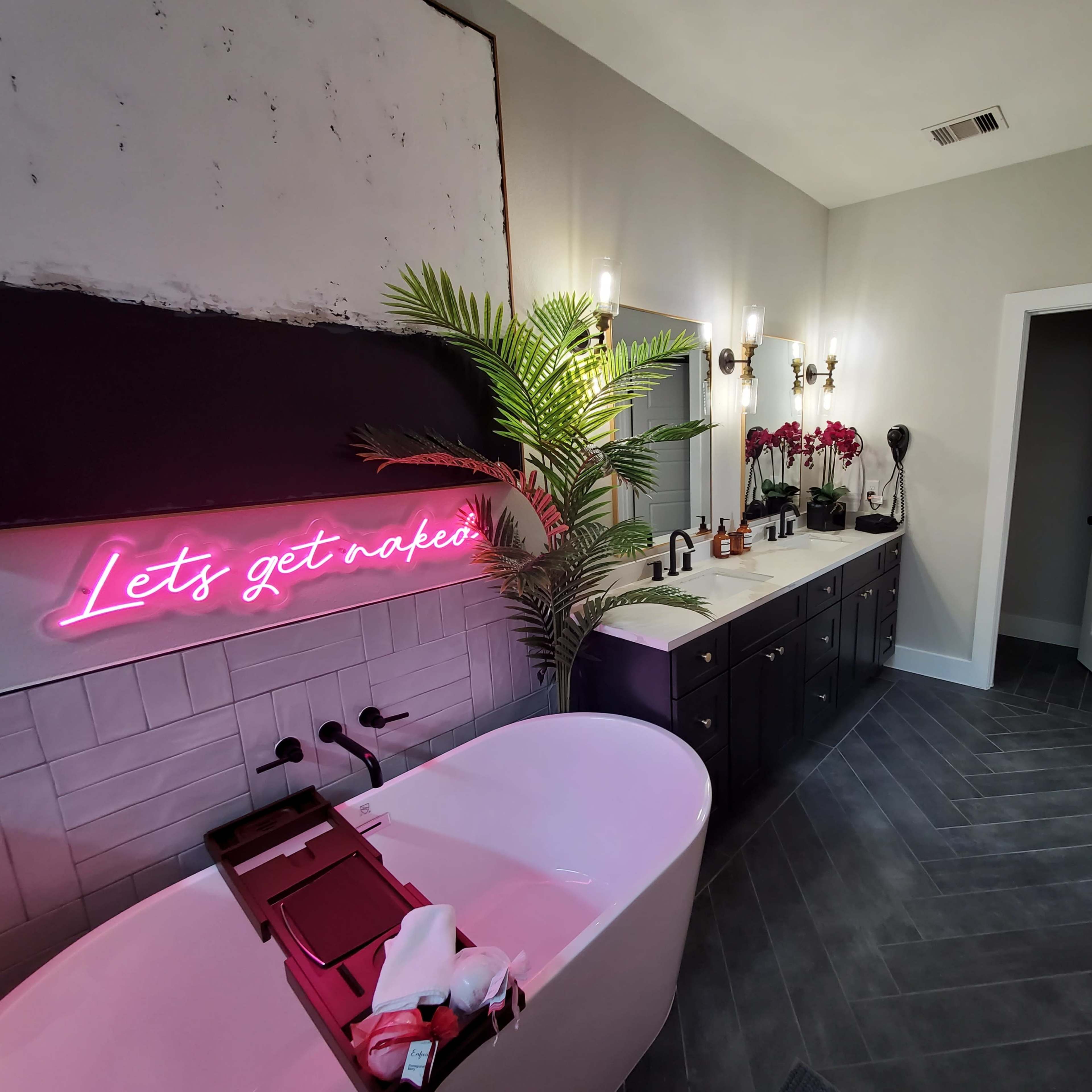 A modern bathroom features a freestanding bathtub, dual sinks with a dark vanity, and a neon sign with the phrase "Let's get naked."