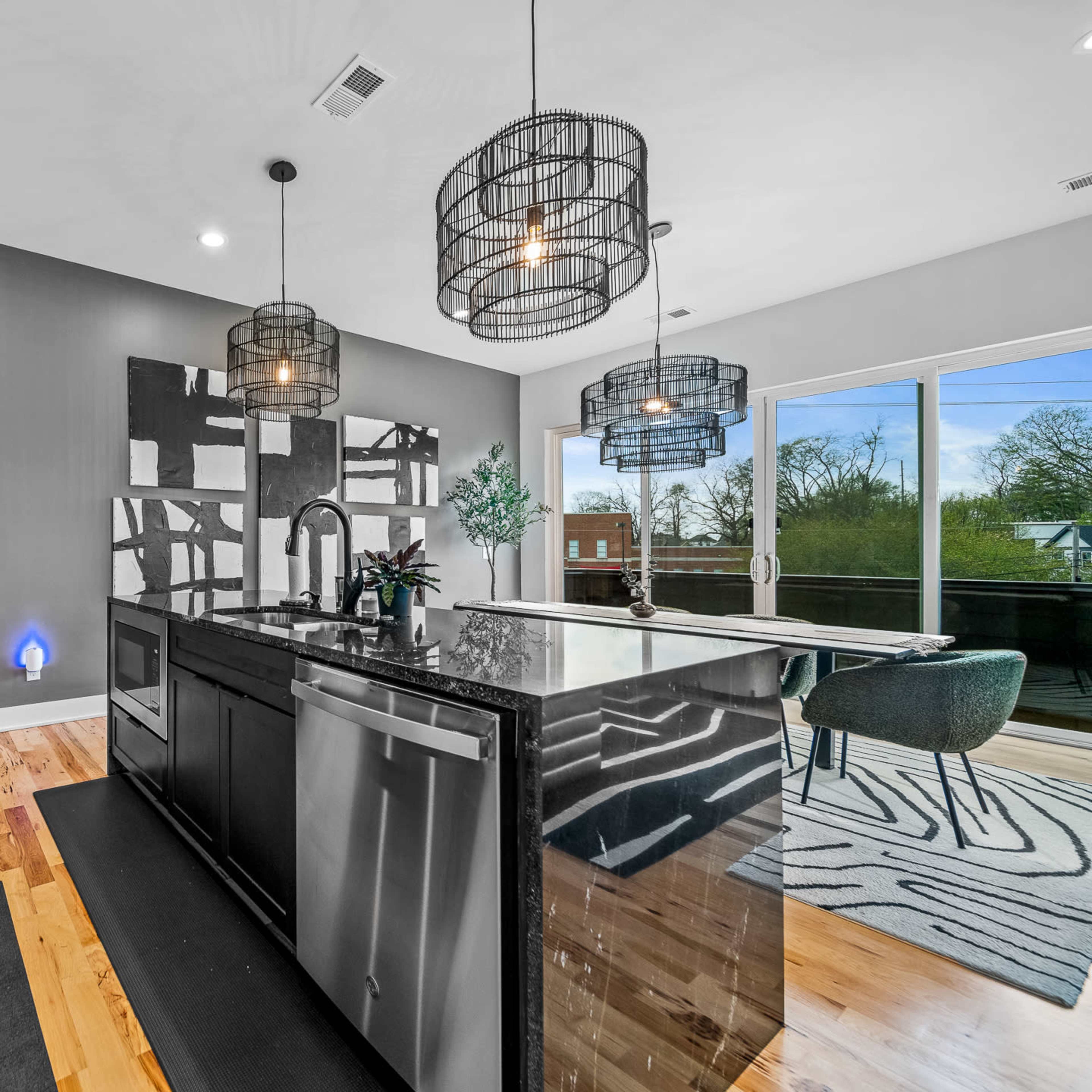 A modern kitchen features a central island with a black countertop, sleek appliances, and large windows overlooking a deck.