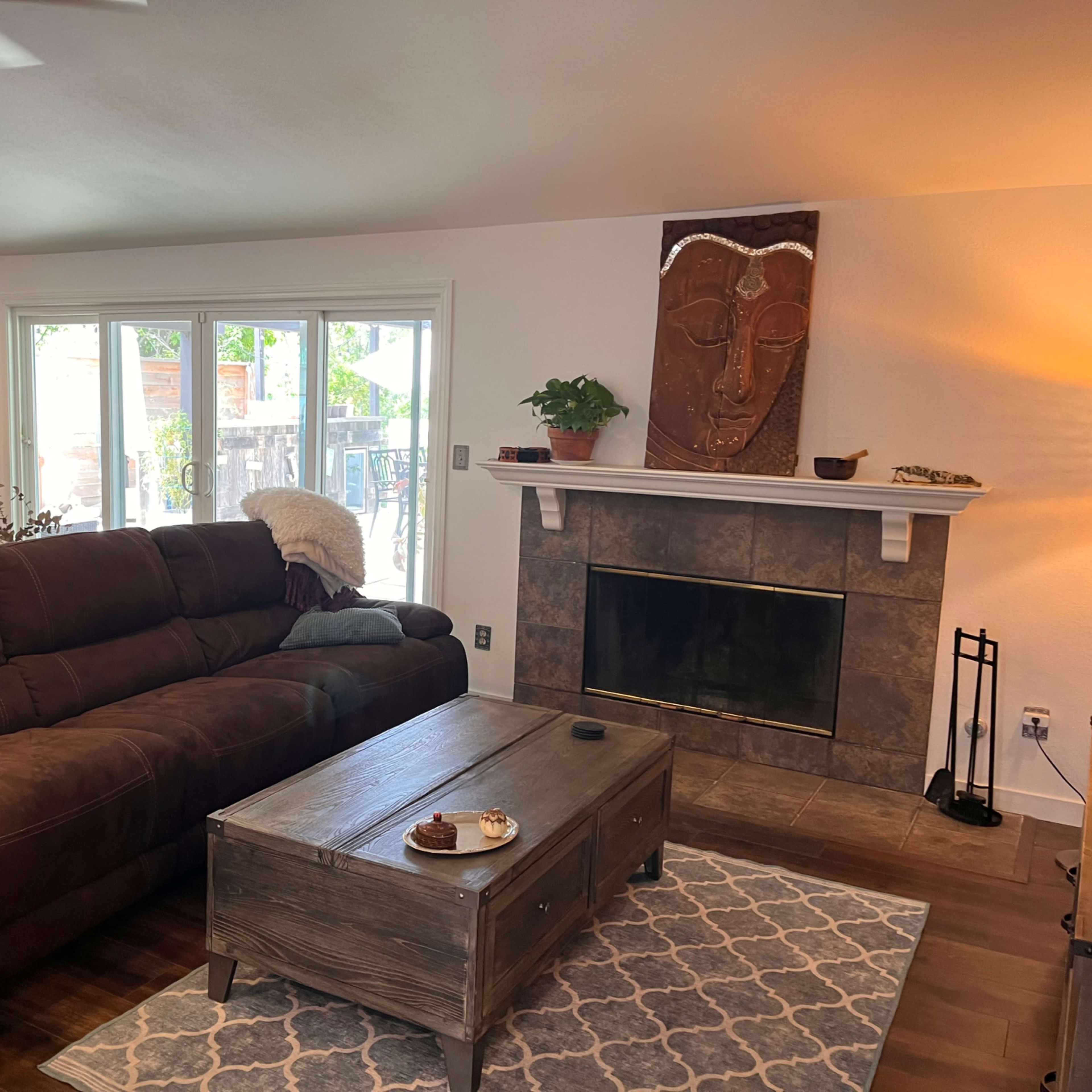 A living room featuring a brown sectional sofa, a wooden coffee table, and a carved wall hanging above a fireplace, with a dining area visible in the background.