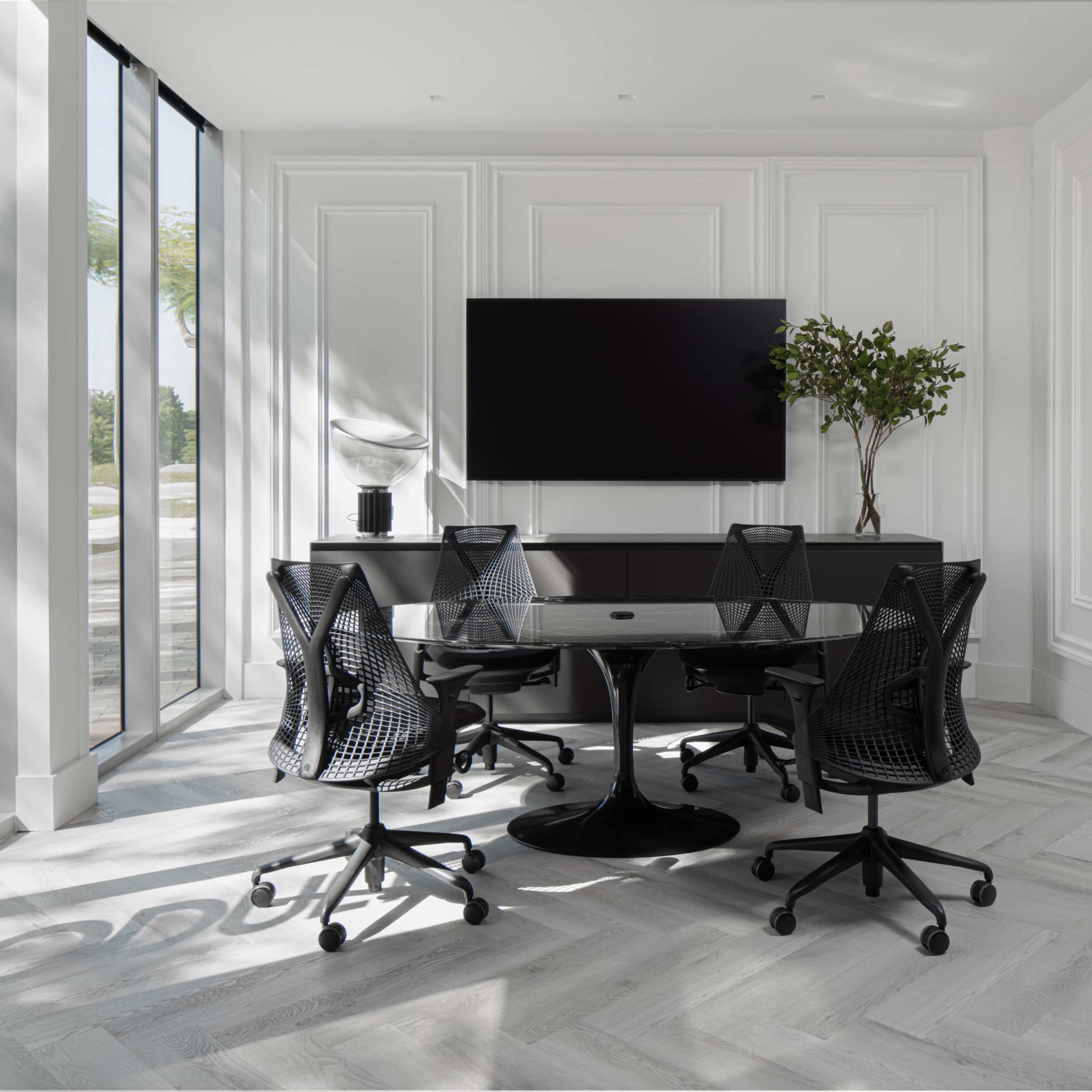 A modern conference room featuring a round black table surrounded by four black office chairs, a large screen on the wall, and a decorative plant on a side cabinet.