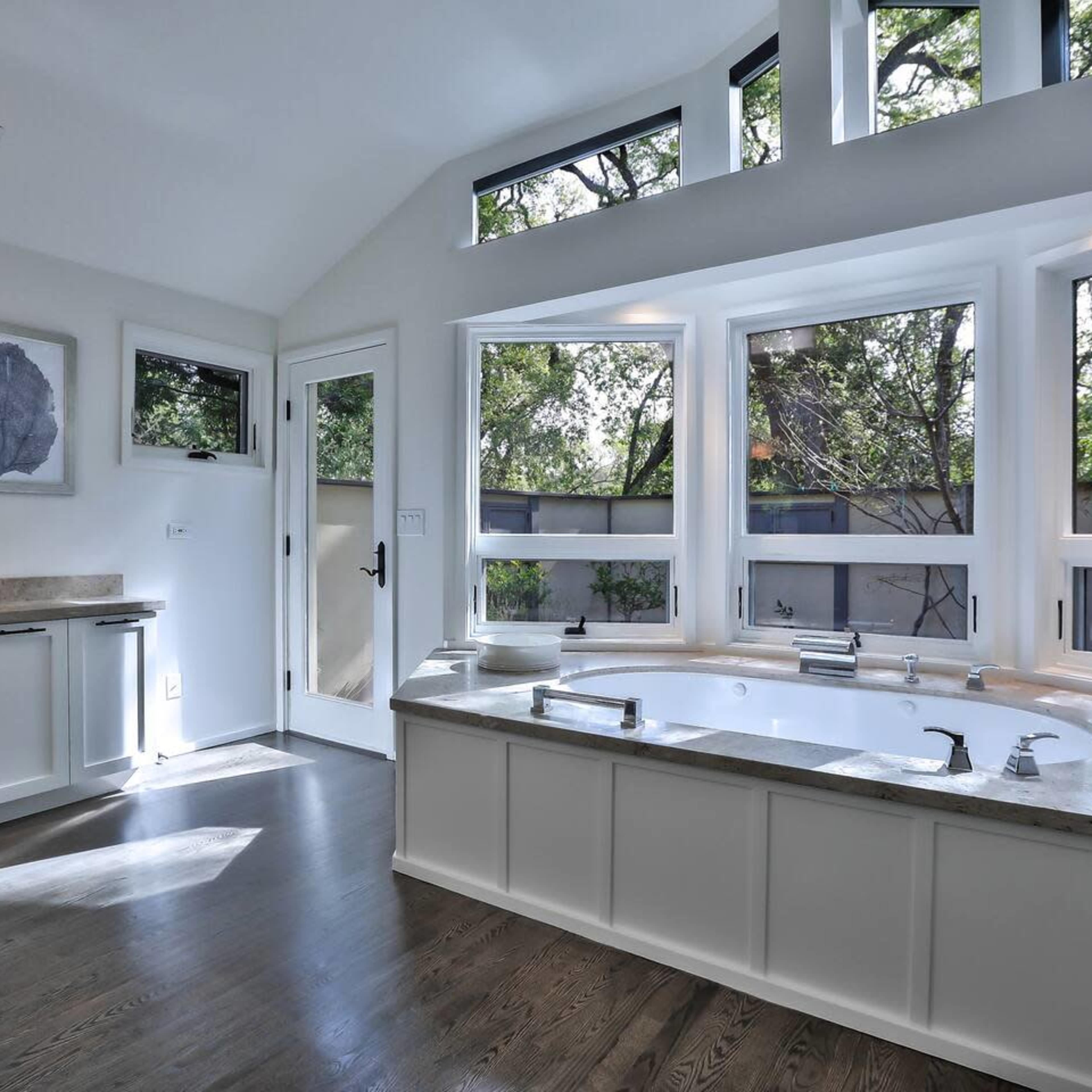 A modern bathroom with a large bathtub positioned under windows that provide natural light and views of greenery outside.