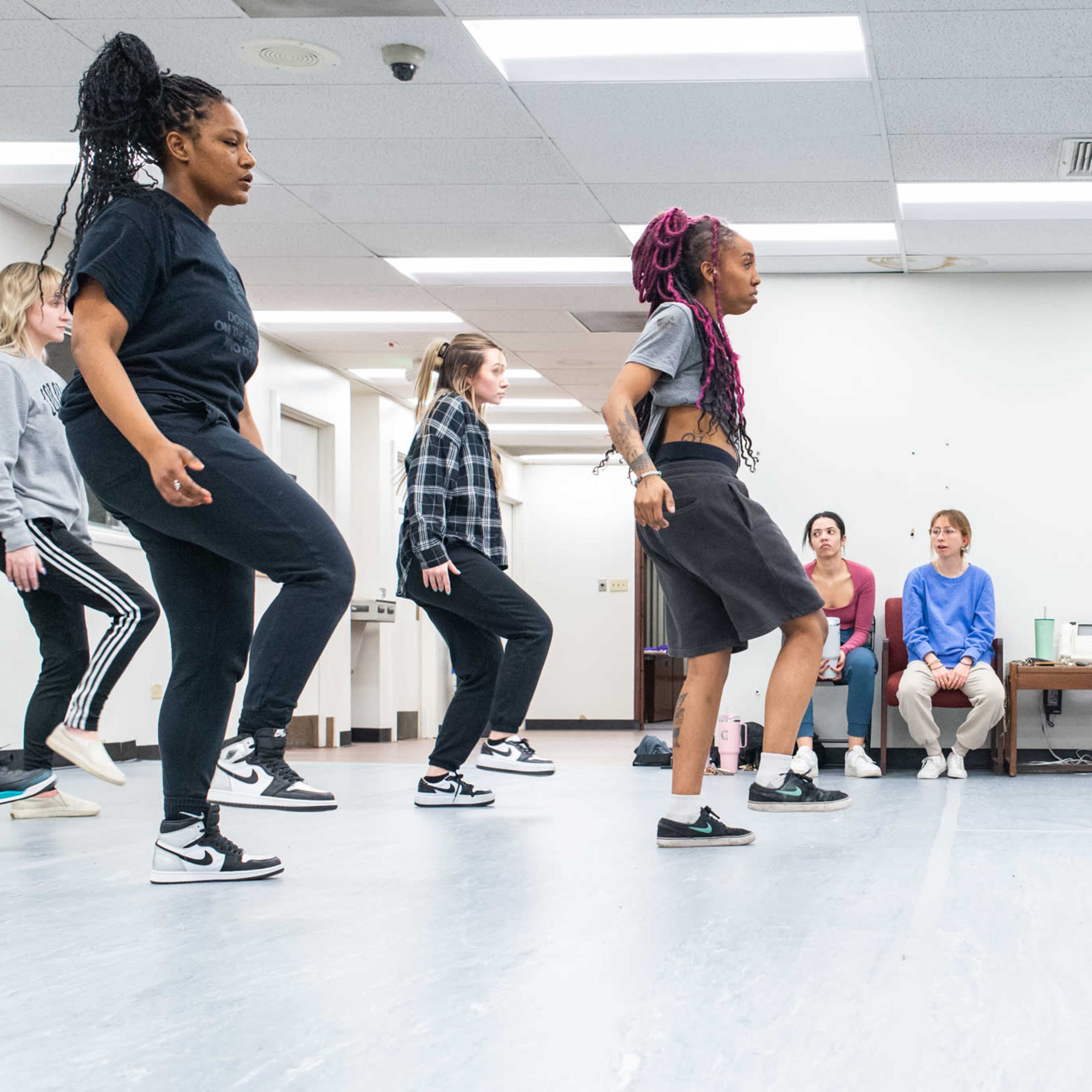 A group of dancers practices footwork in a spacious studio while several observers sit on the side.