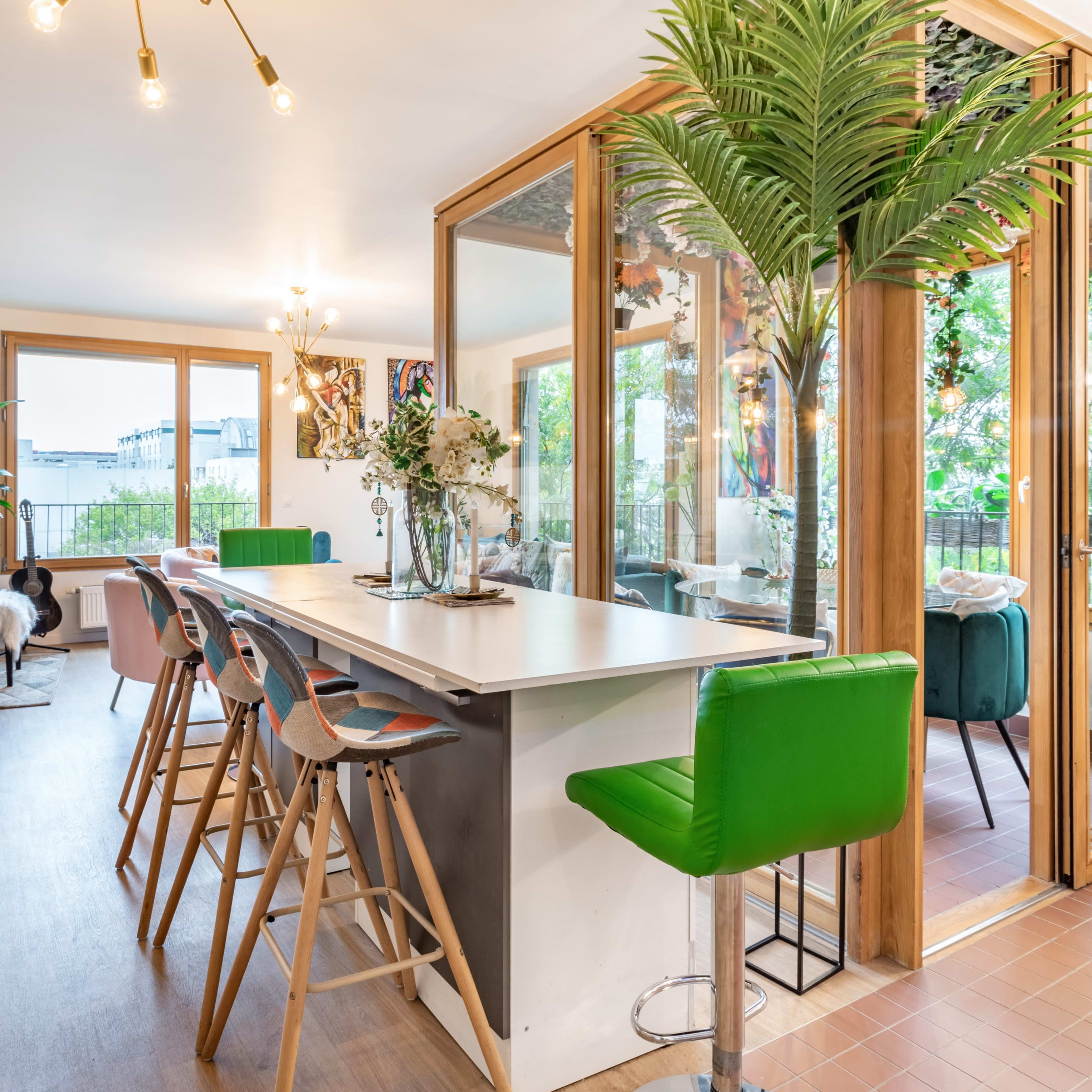 A modern open-concept living space features a kitchen island with bar stools, a dining area, and a glass-enclosed room filled with plants.