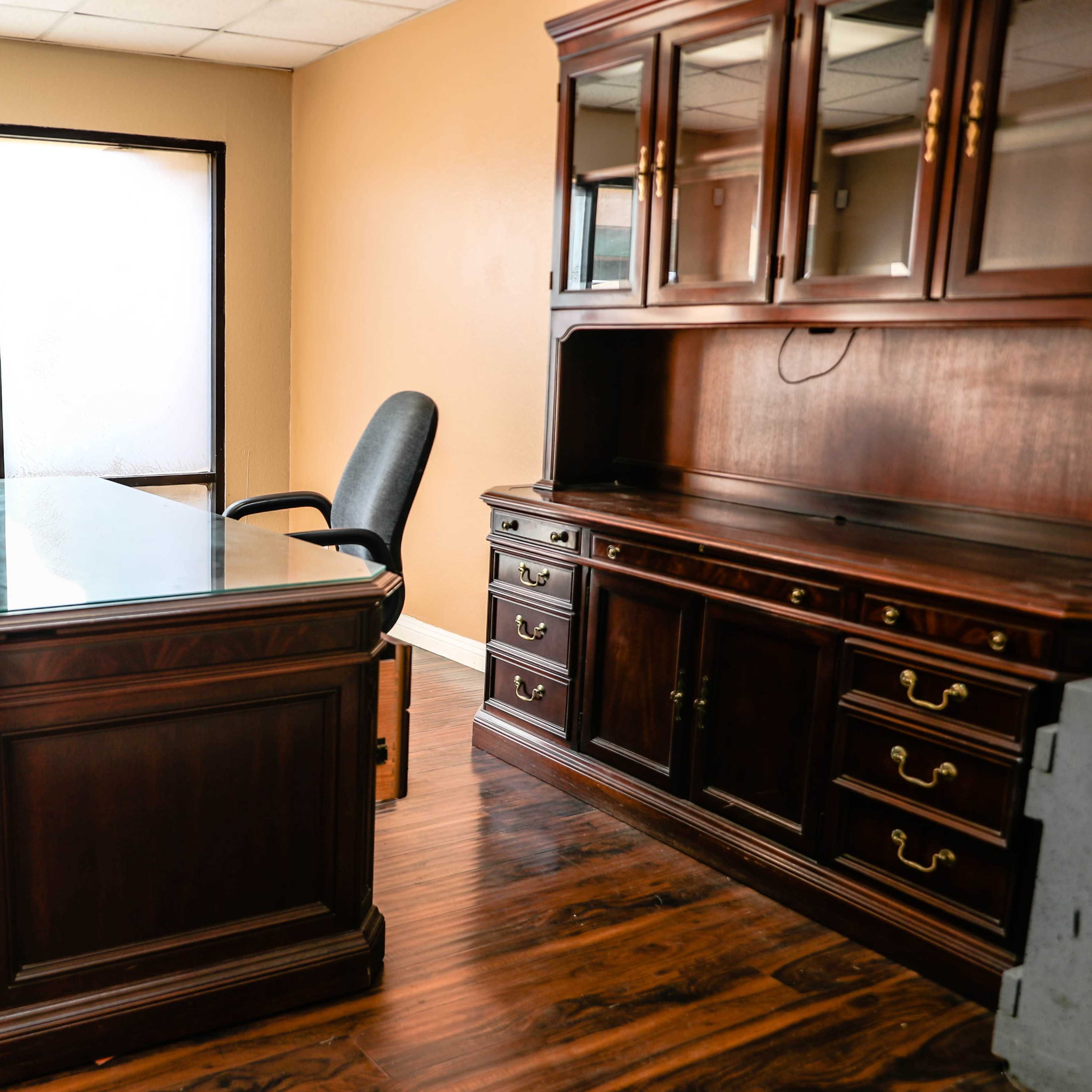 A spacious office with a wooden desk, an ergonomic chair, and a matching wooden cabinet, next to a large window.