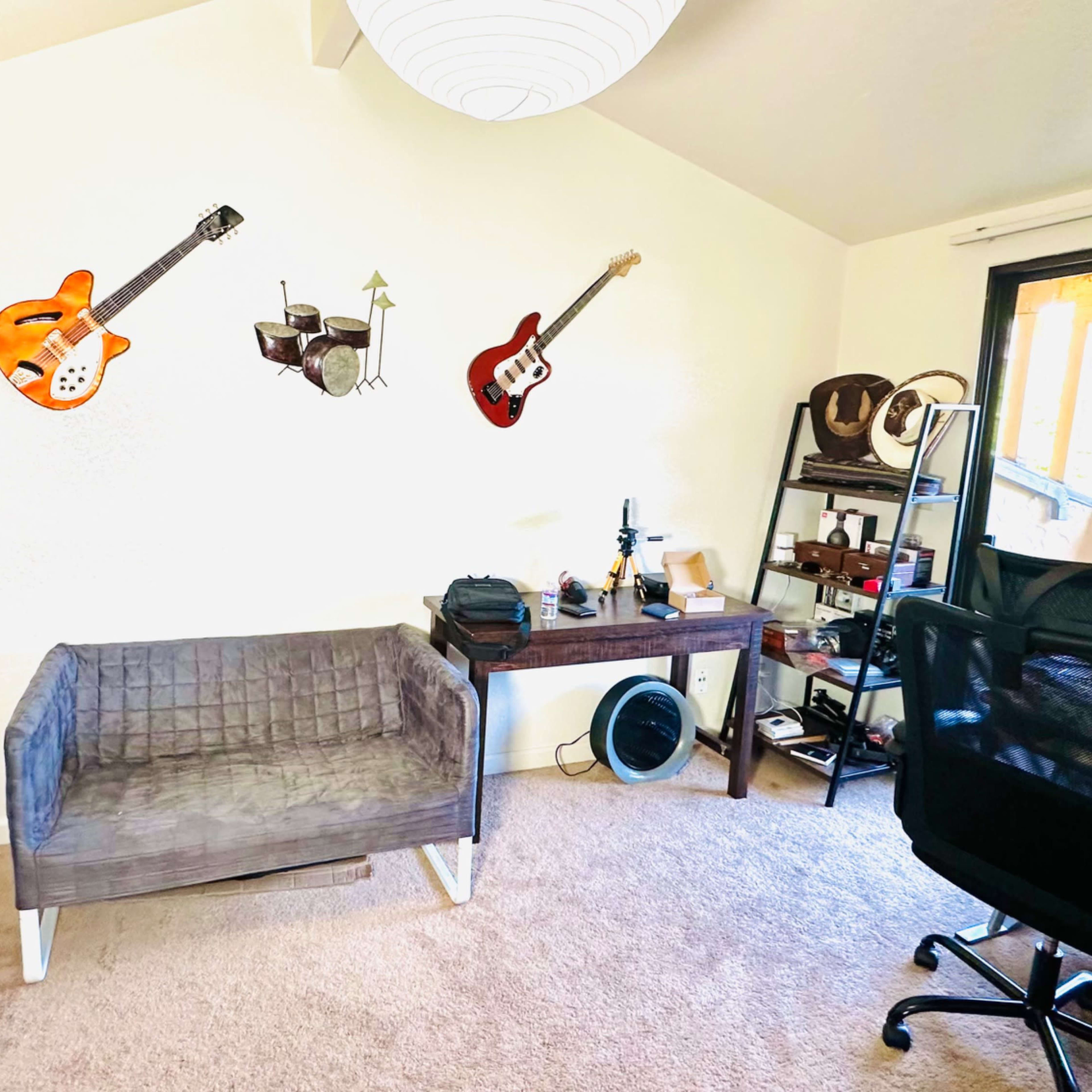 The room features a gray couch, a desk with various items, a black office chair, and three guitars mounted on the wall.