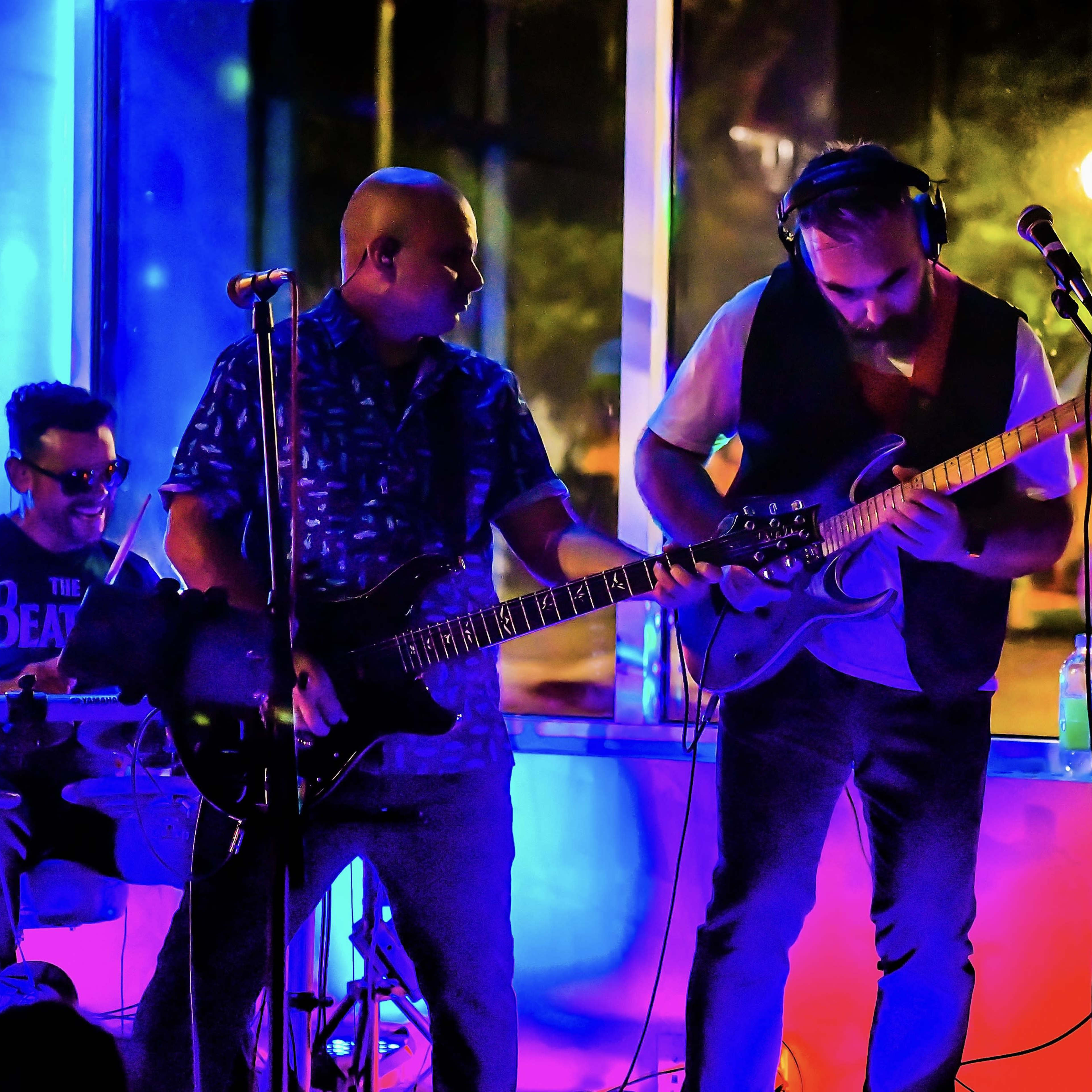 A band performs on stage with a drummer, guitarist, and singer under colorful lighting.