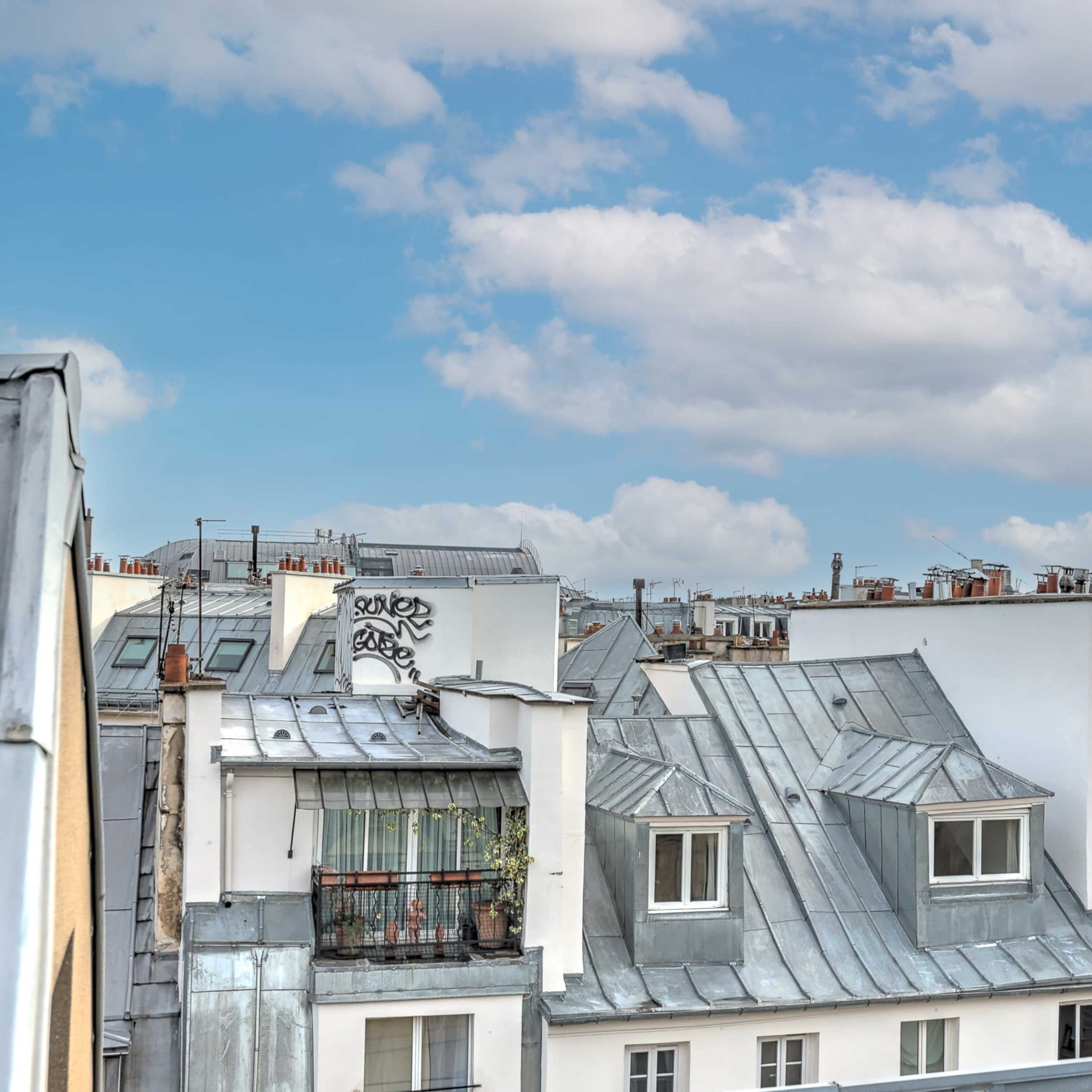 A view of rooftops in a city, featuring a mix of metal and tiled roofs, balconies, and scattered graffiti under a partly cloudy sky.