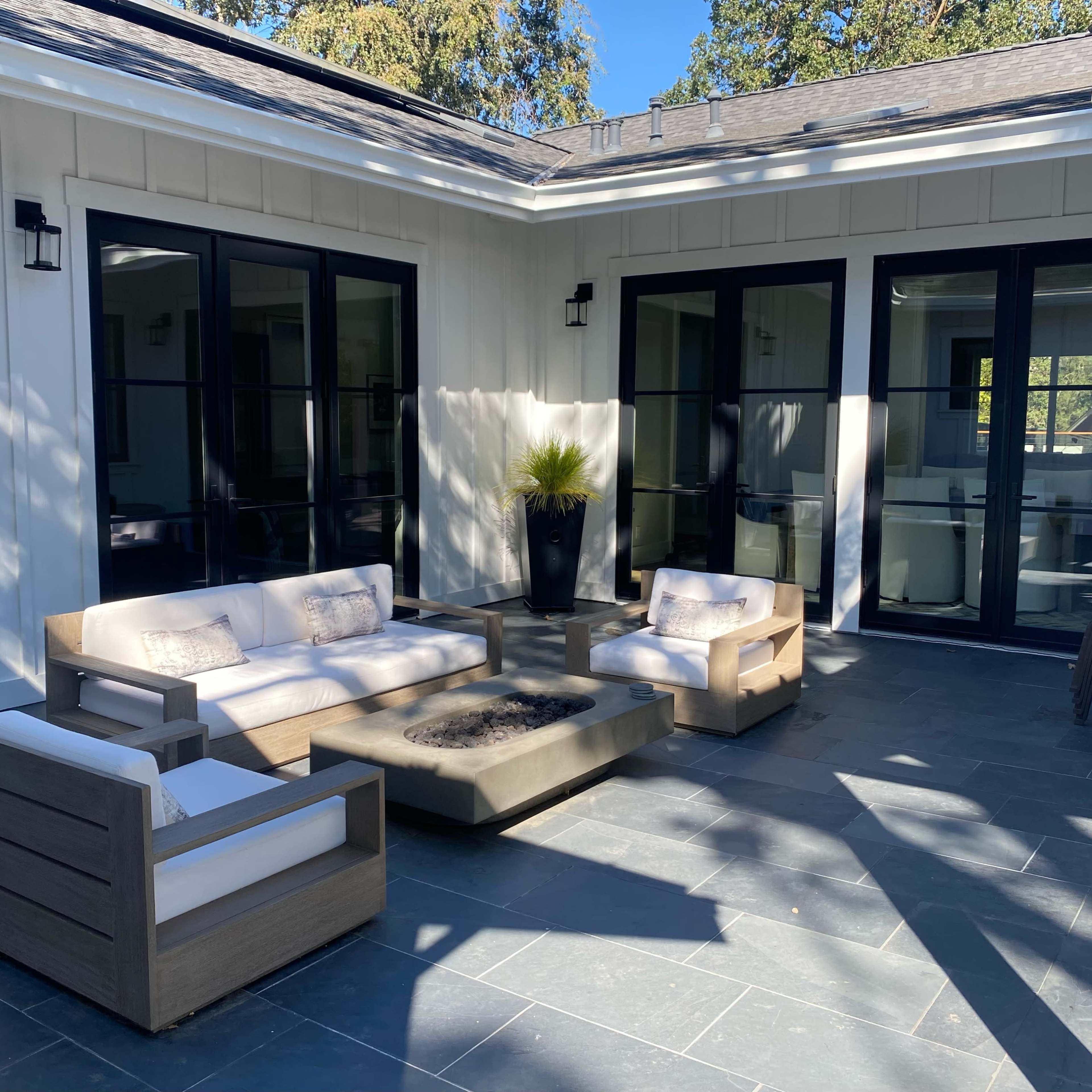 A modern patio features a seating area with beige sofas and a coffee table, surrounded by large windows and a potted plant.