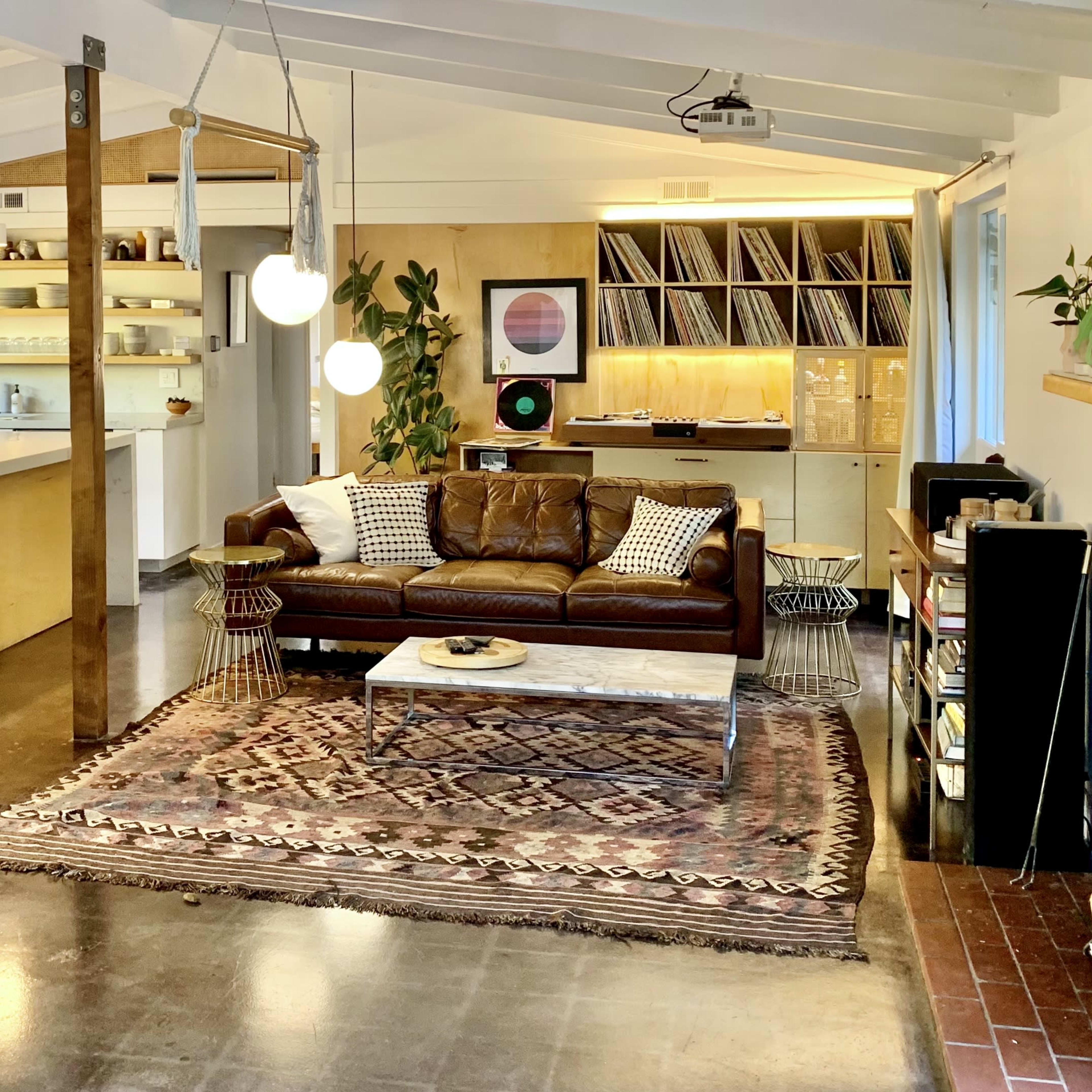 A modern indoor living space with a brown leather sofa, a large coffee table, an area rug, and a minimalist kitchen featuring open shelving and a wall of records.
