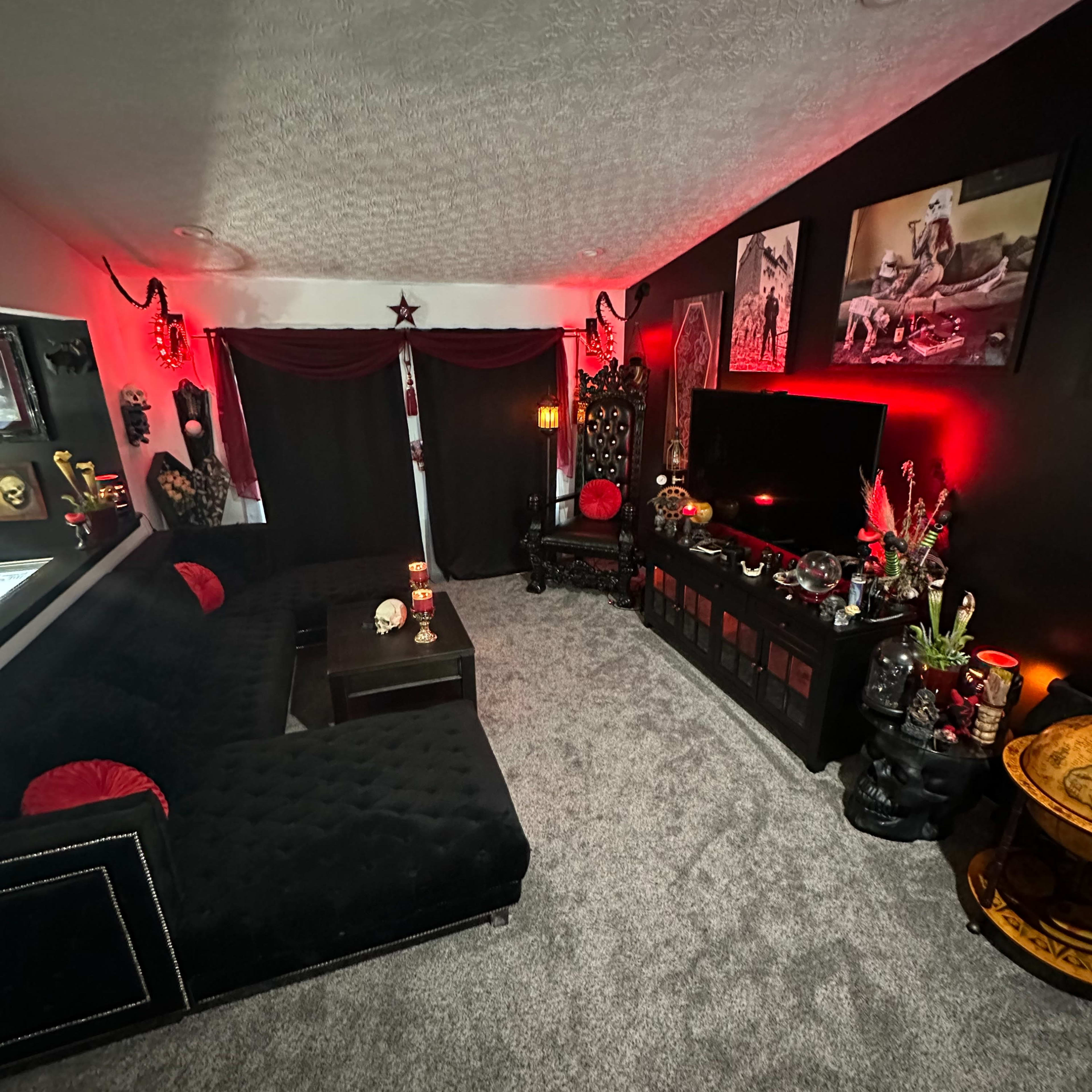 A dark-themed living room with black walls, a plush black couch, and decorative elements including red accents, artwork, and various ornate objects.