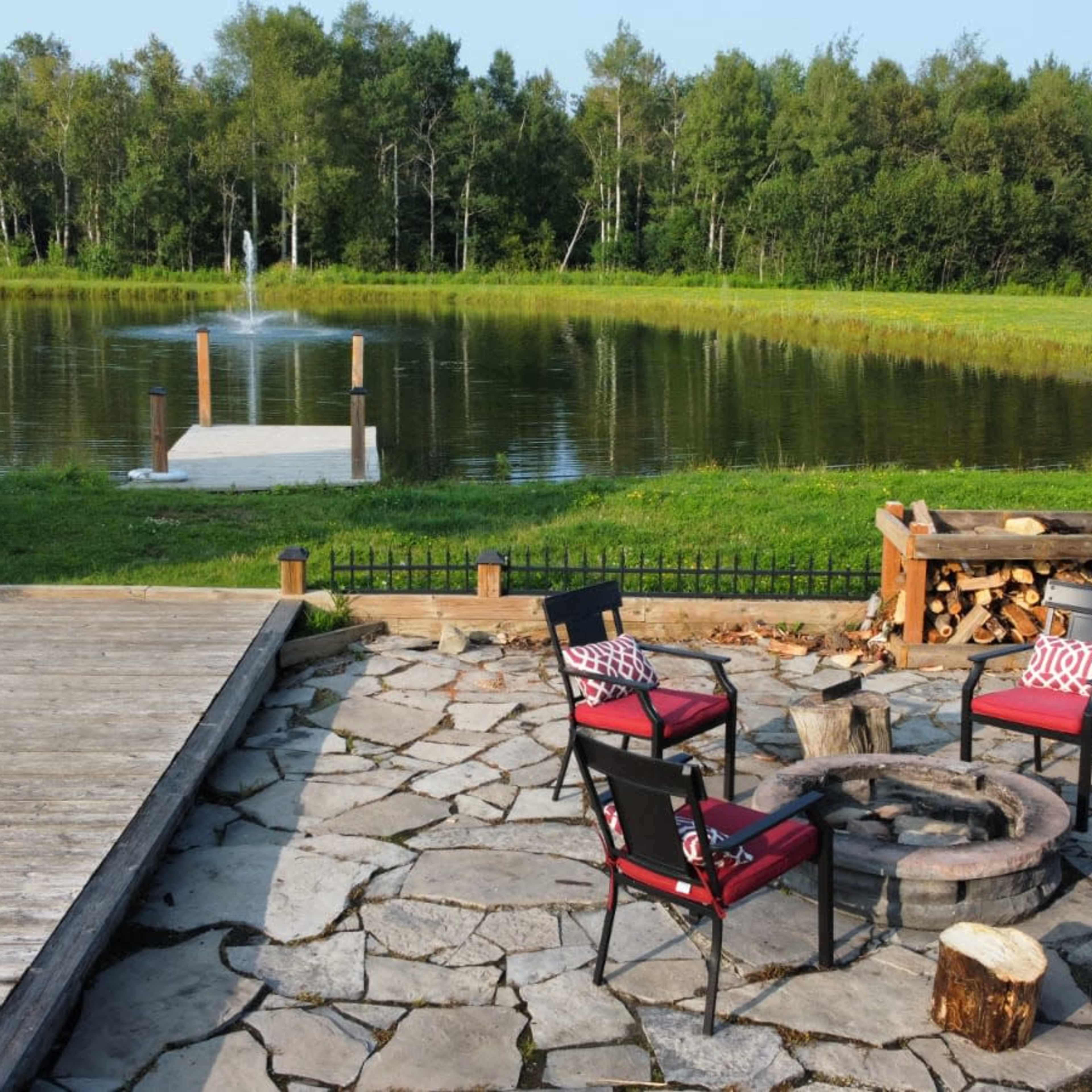 A stone patio with four chairs arranged around a fire pit, situated beside a pond with a fountain and a small dock.