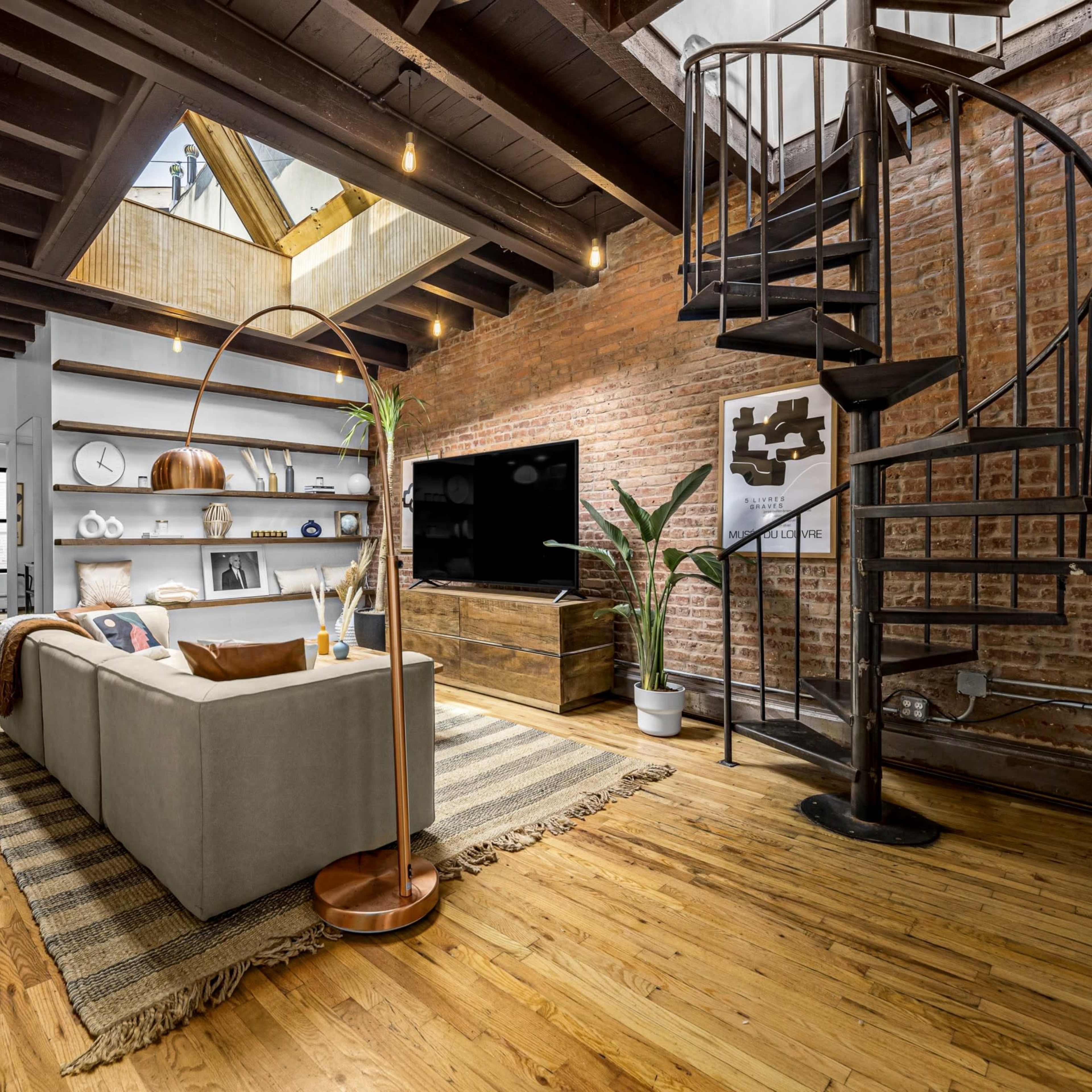 A modern living room with a spiral staircase, exposed brick walls, wooden beams, and a large sectional sofa facing a television mounted on the wall.