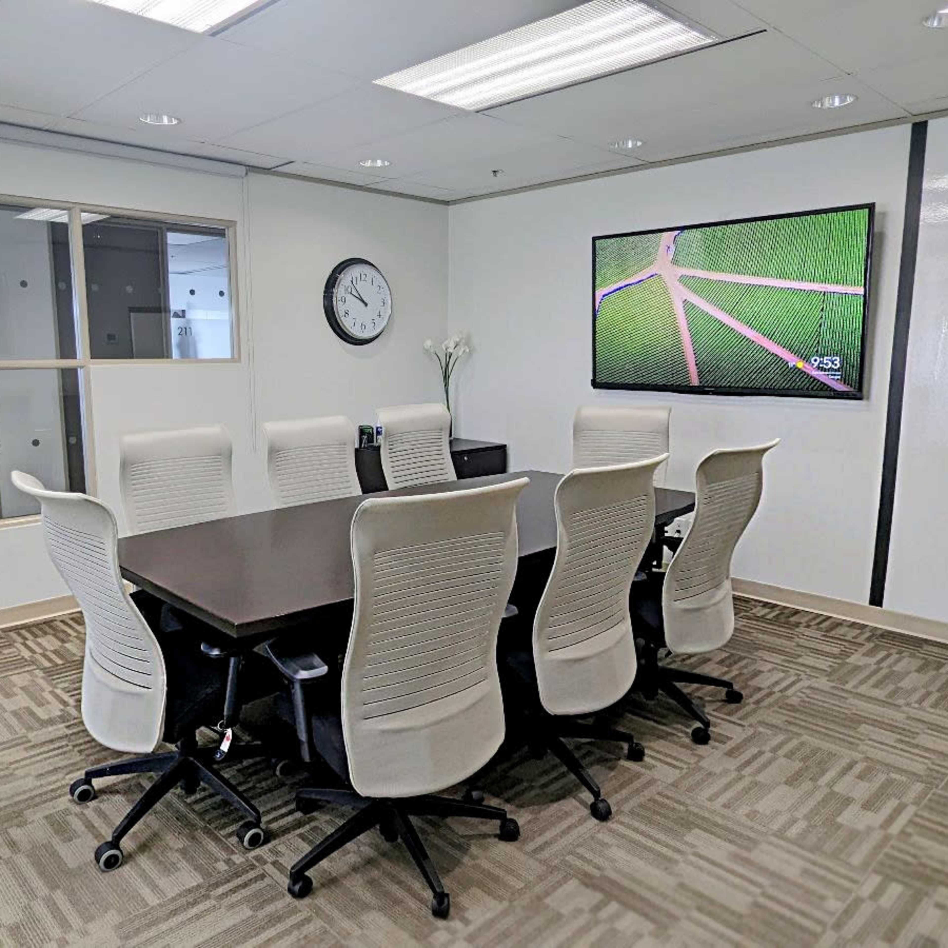 A modern conference room features a large table surrounded by seven white chairs, a wall-mounted screen displaying green fields, and a clock on the wall.