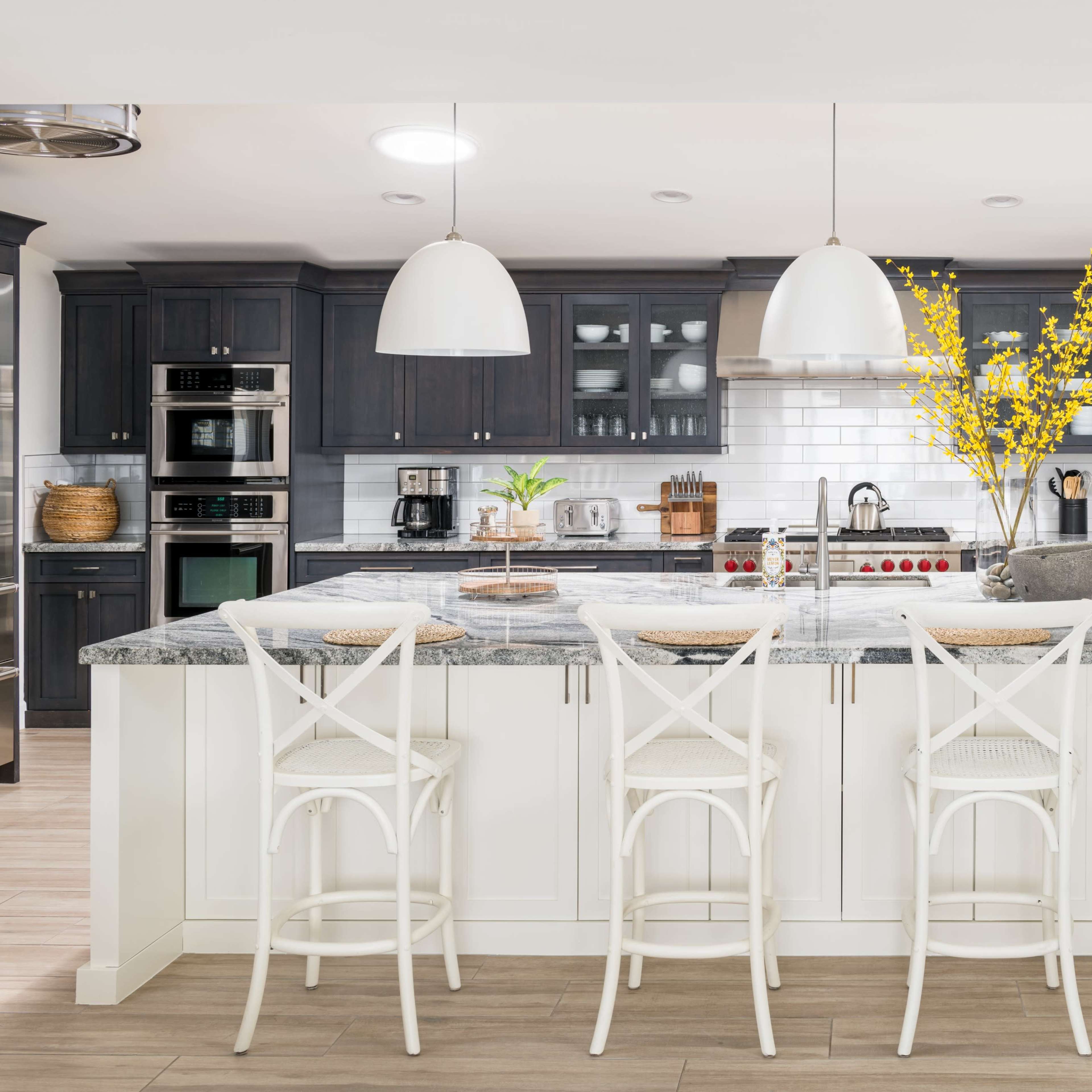A modern kitchen features a large island with four white stools, dark cabinetry, stainless steel appliances, and decorative yellow flowers in a central vase.