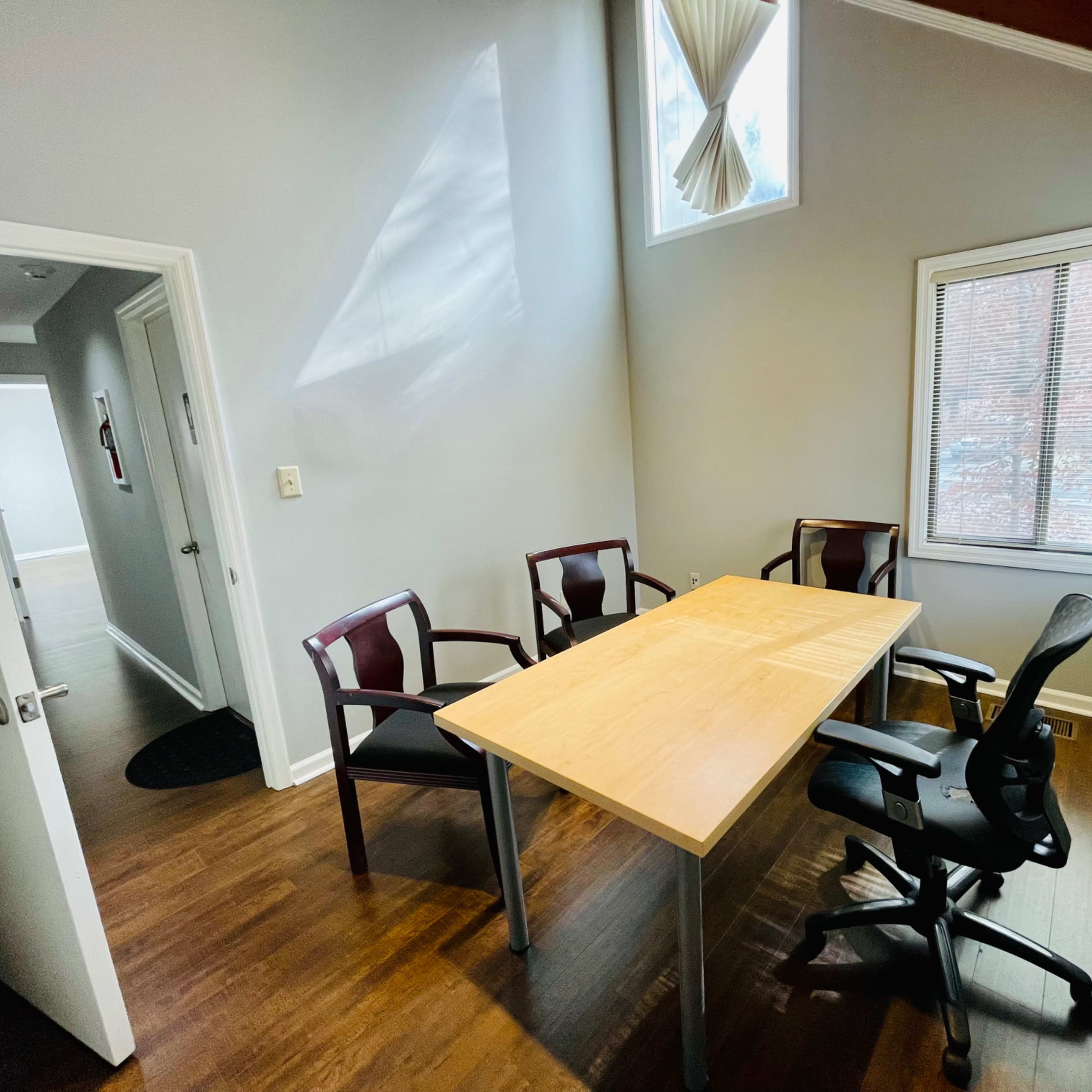A small meeting room with a wooden table and four chairs, featuring a window with natural light.
