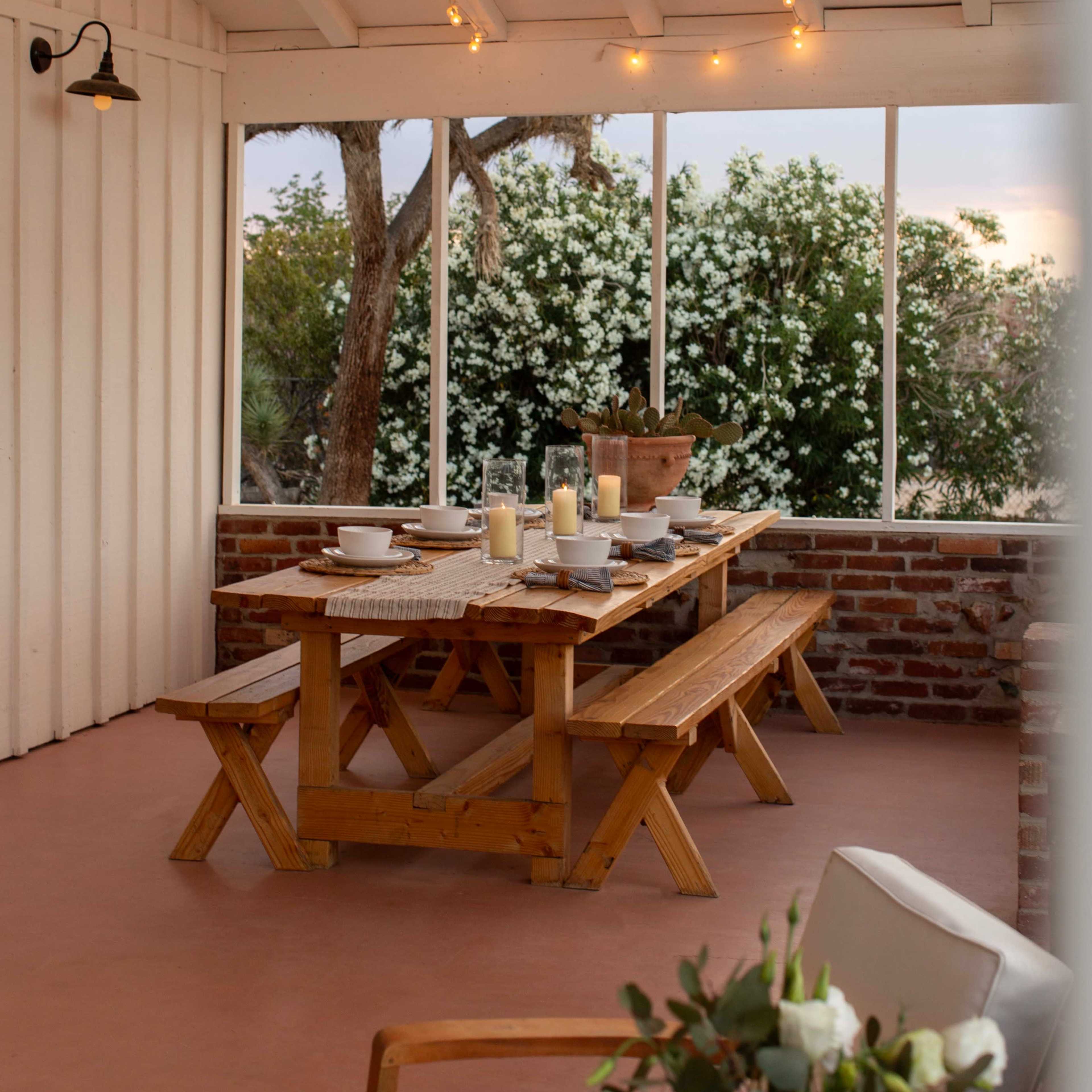 A wooden dining table is set with dishes and candles in a bright, airy room with large windows overlooking flowering plants.