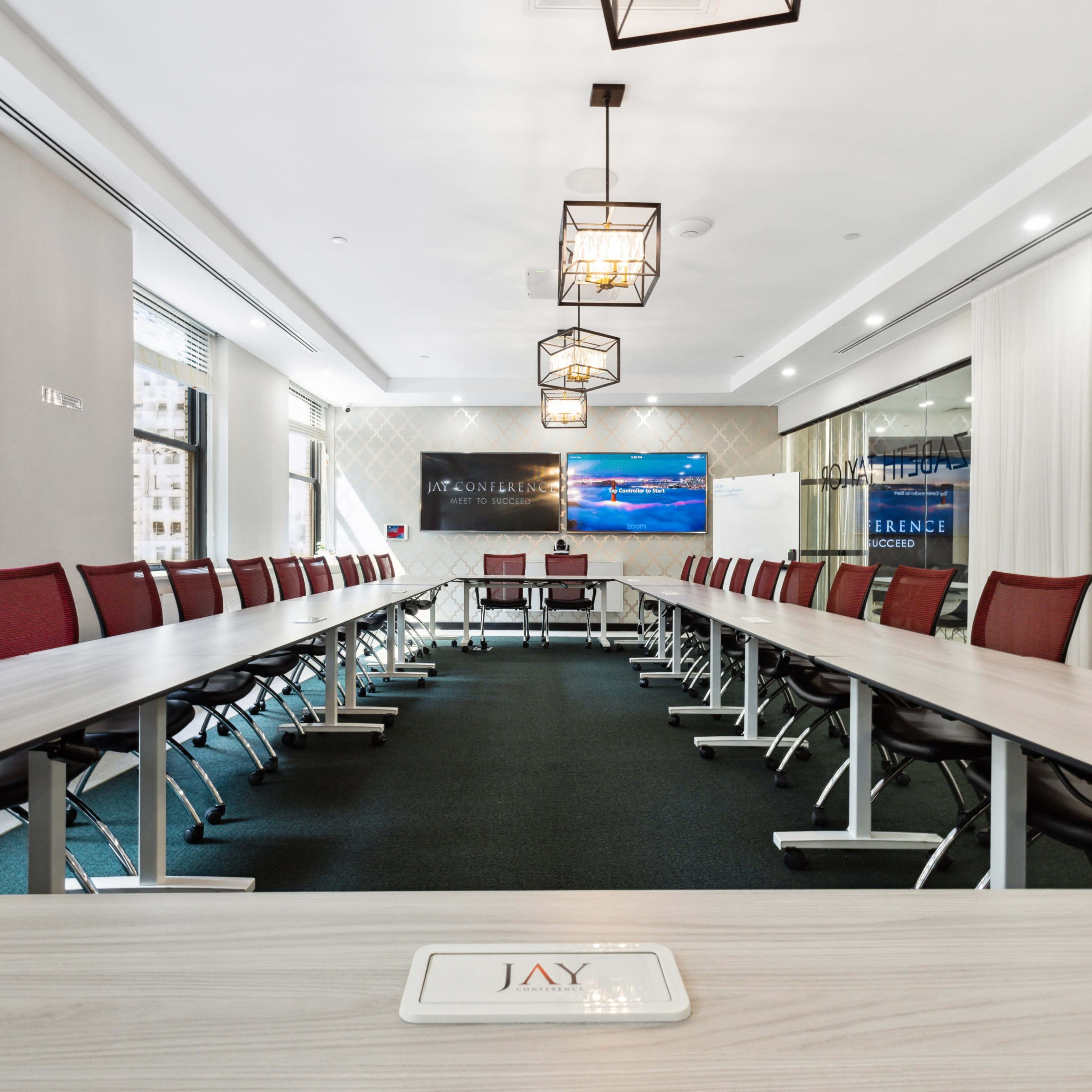 A spacious conference room with a long rectangular table, surrounding red chairs, and a presentation screen at one end.