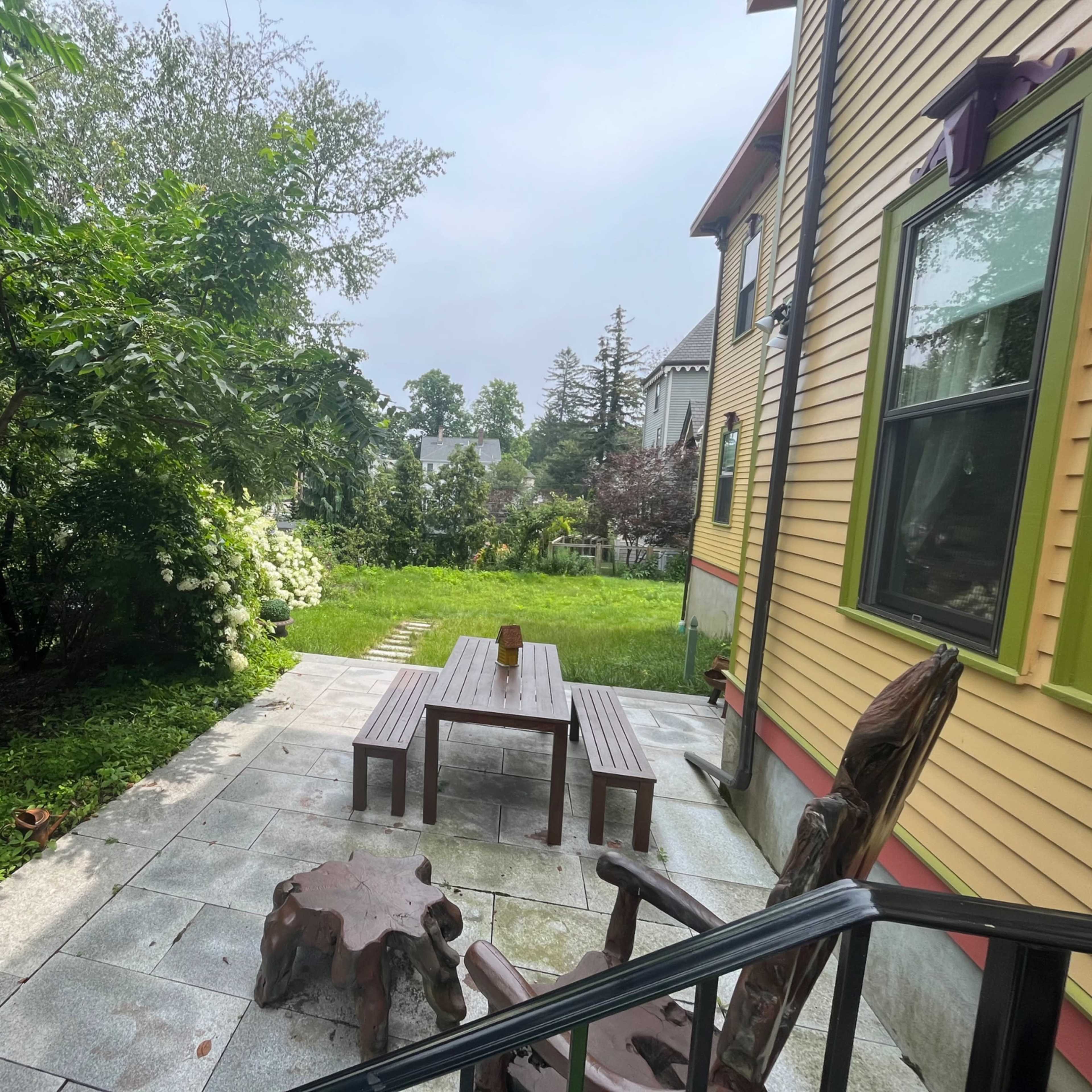 A stone patio features a wooden table and benches, surrounded by lush greenery and a well-maintained lawn.