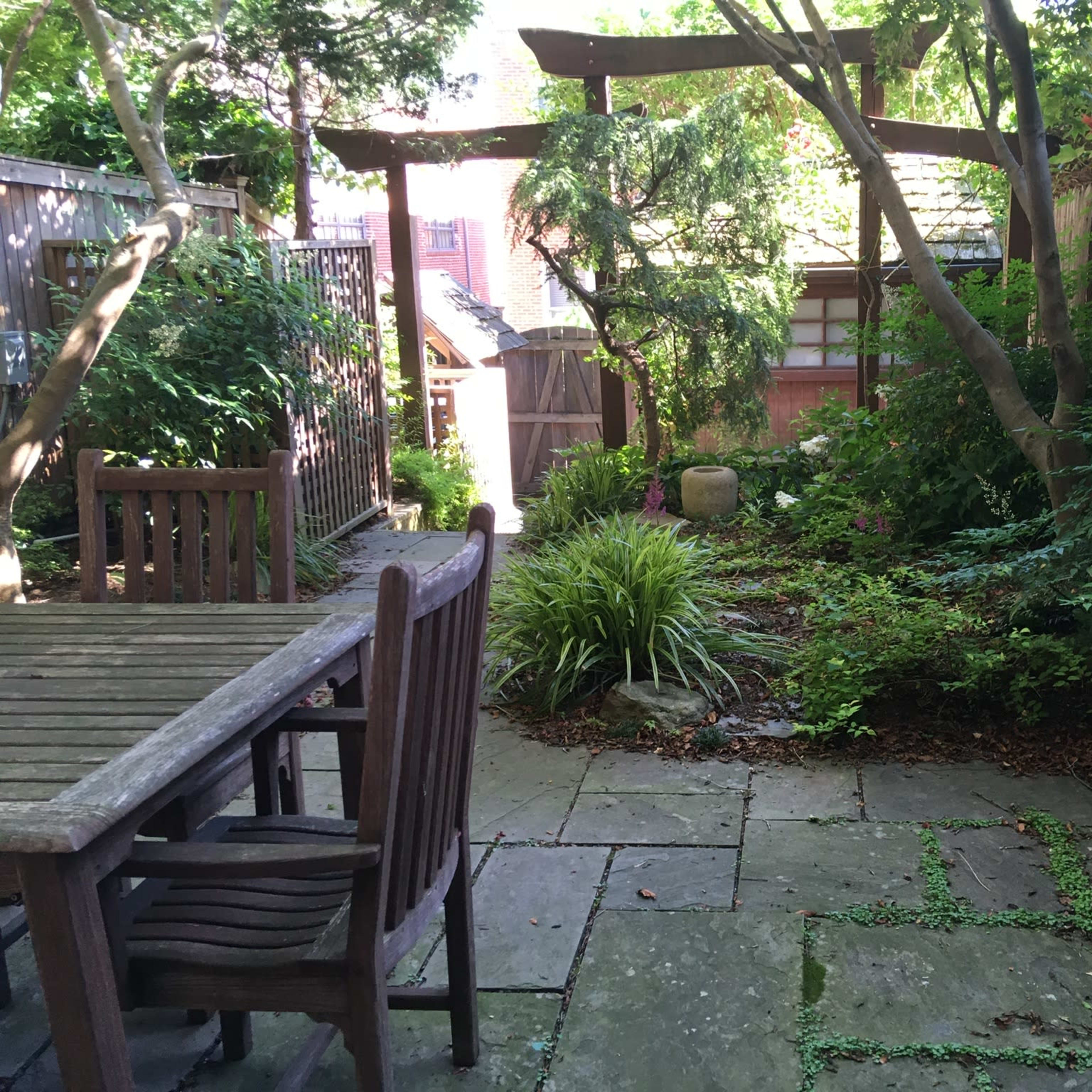 A stone-paved garden area with wooden furniture, surrounded by greenery and a trellis overhead.