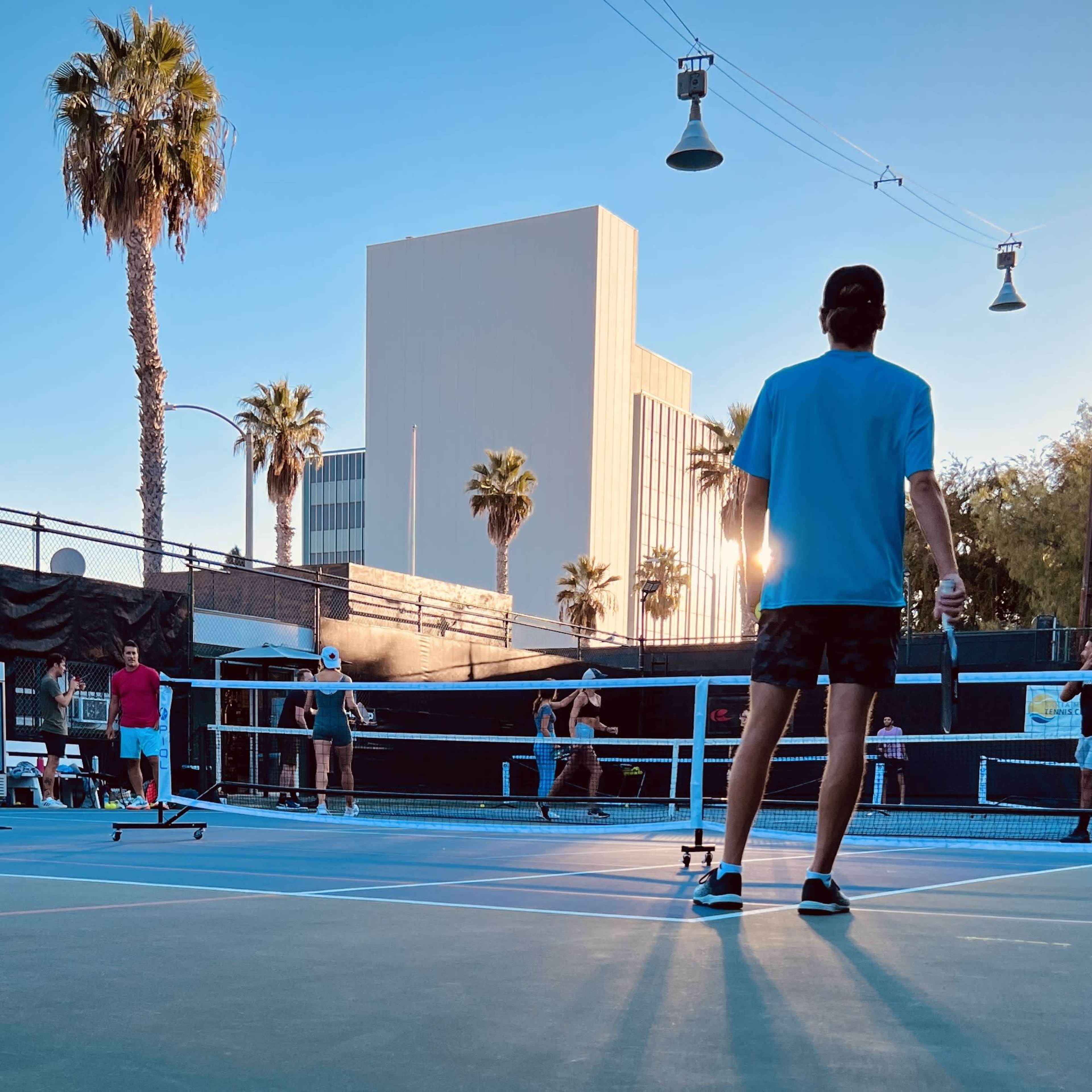 A group of people play tennis on outdoor courts against a backdrop of palm trees and a tall building during sunset.