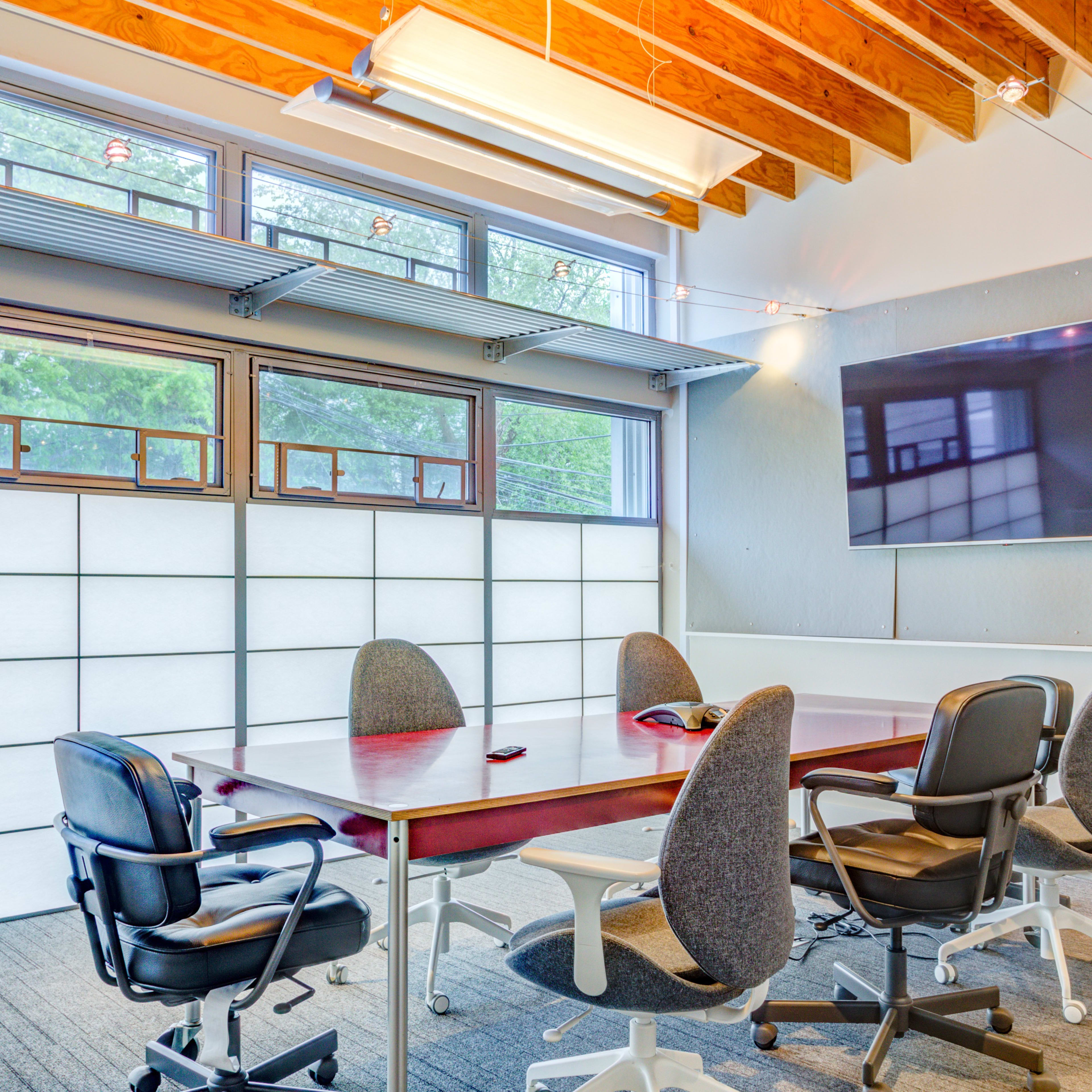 A modern conference room features a large table surrounded by rolling chairs, large windows, and a screen mounted on the wall.