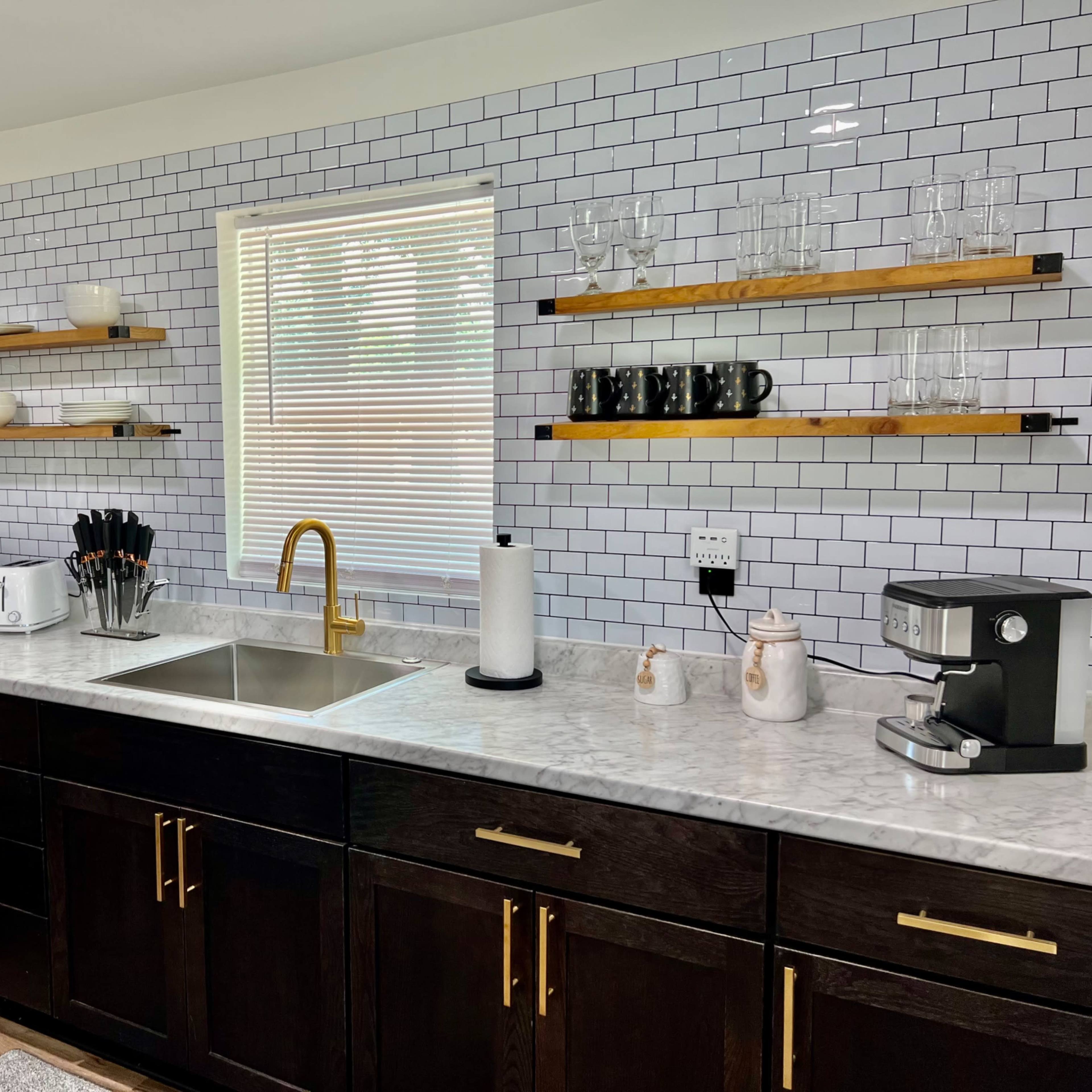 A modern kitchen with dark cabinets, a marble countertop, open shelving, and various appliances including a microwave, kettle, and coffee machine.