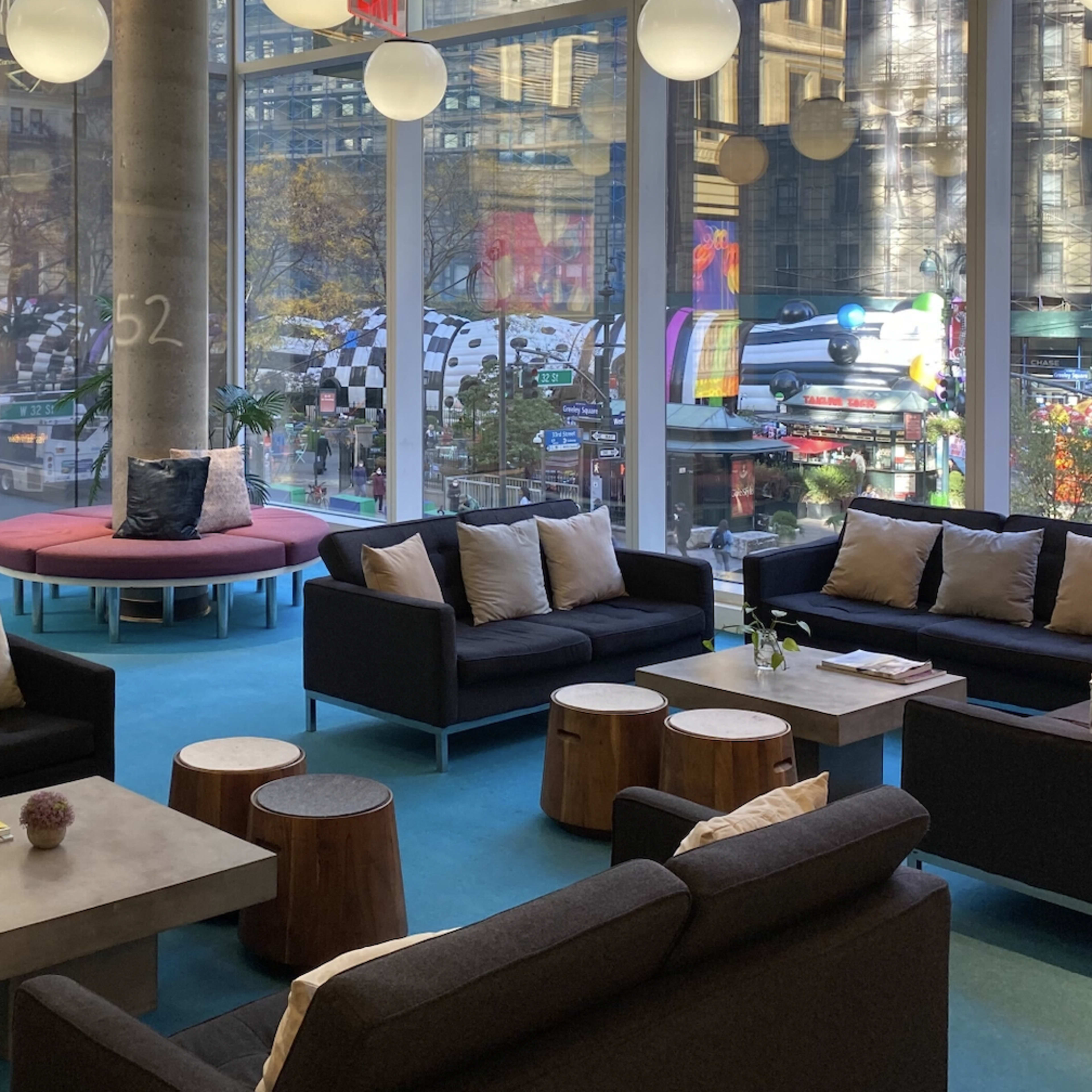 A spacious lounge area with several black sofas and wooden tables, overlooking a busy street filled with colorful decorations.