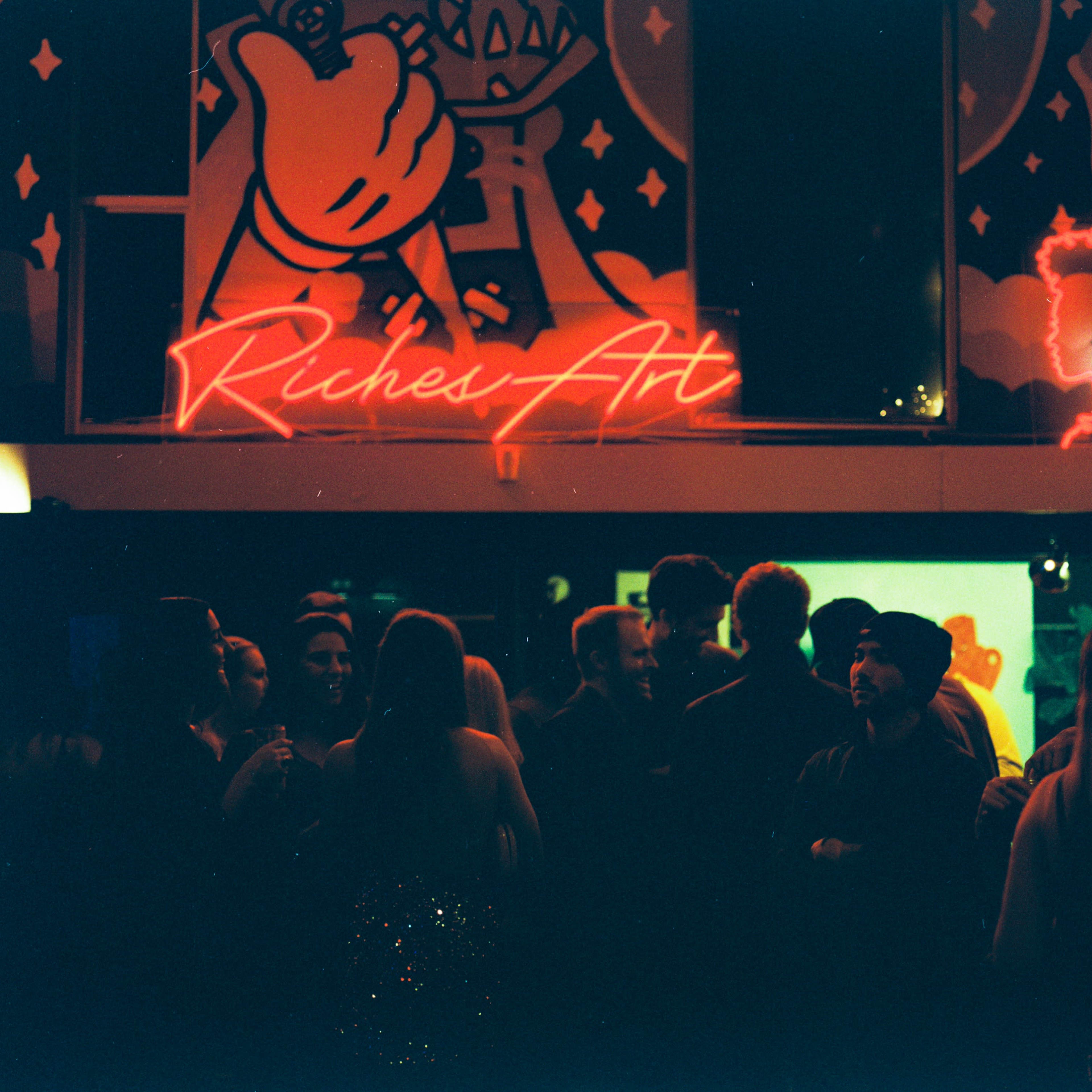 A crowd of people mingles in a dimly lit space featuring vibrant wall art and a glowing neon sign that reads "Riches Art."