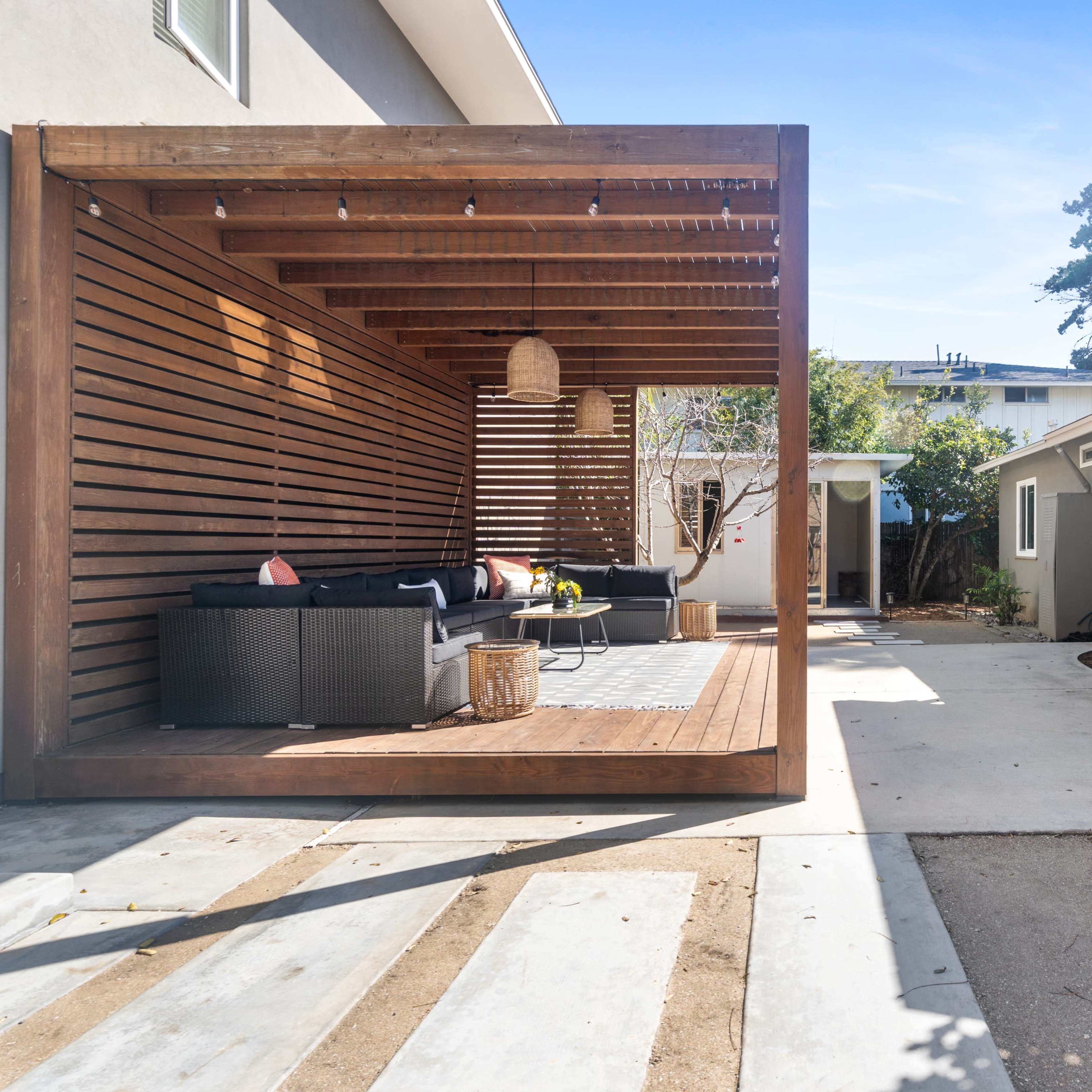A modern outdoor patio area with a wooden pergola, featuring a seating arrangement and a grill beside a house.