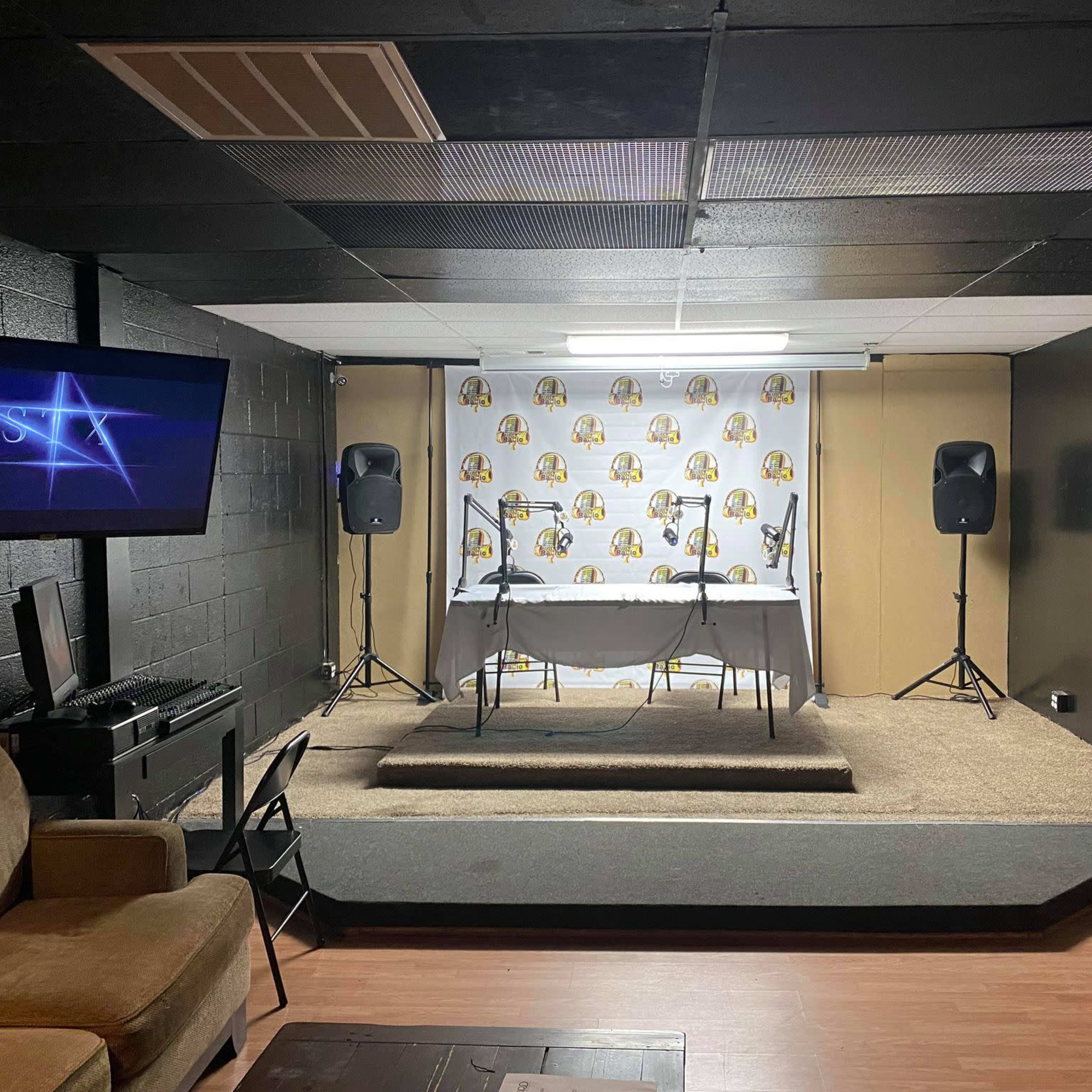A small, stage area set up with a table and two microphones, flanked by speakers, in a room with a black backdrop and a branded backdrop featuring a logo.