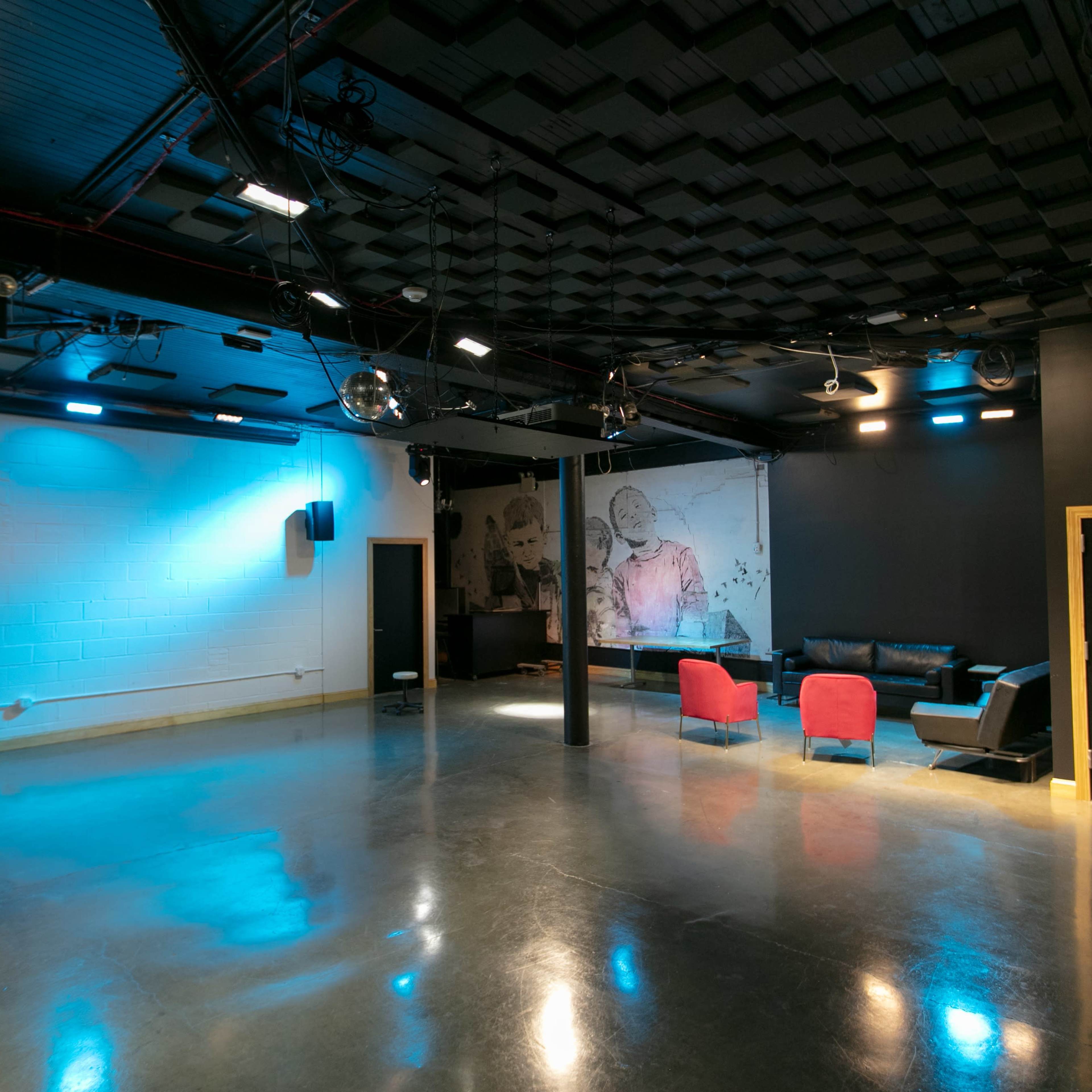 An empty indoor space with polished concrete floors, black walls, a large mural, and seating areas featuring a dark couch and red chairs.