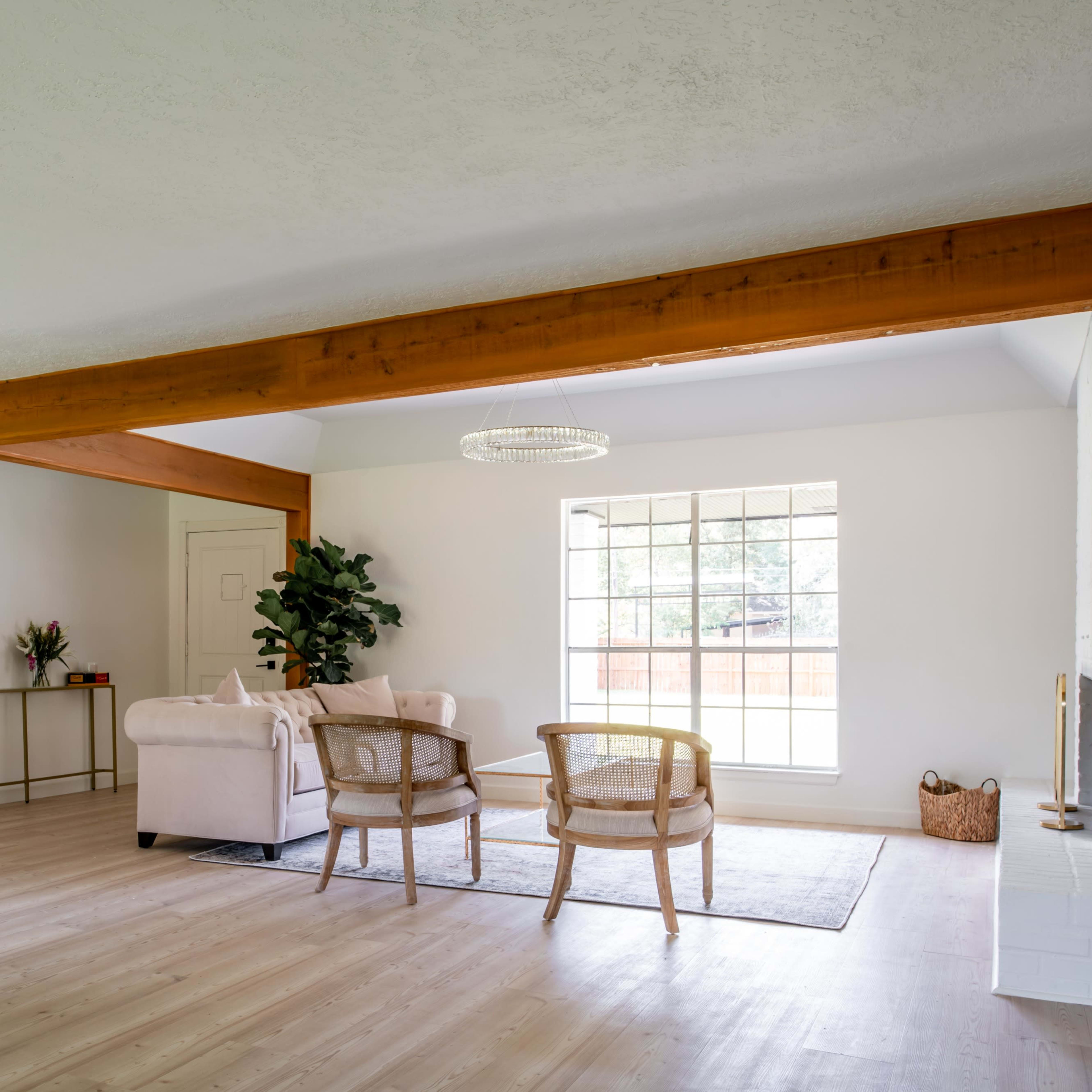 A bright living room features a beige sofa, two wooden chairs, and a large window, with exposed wooden beams on the ceiling.
