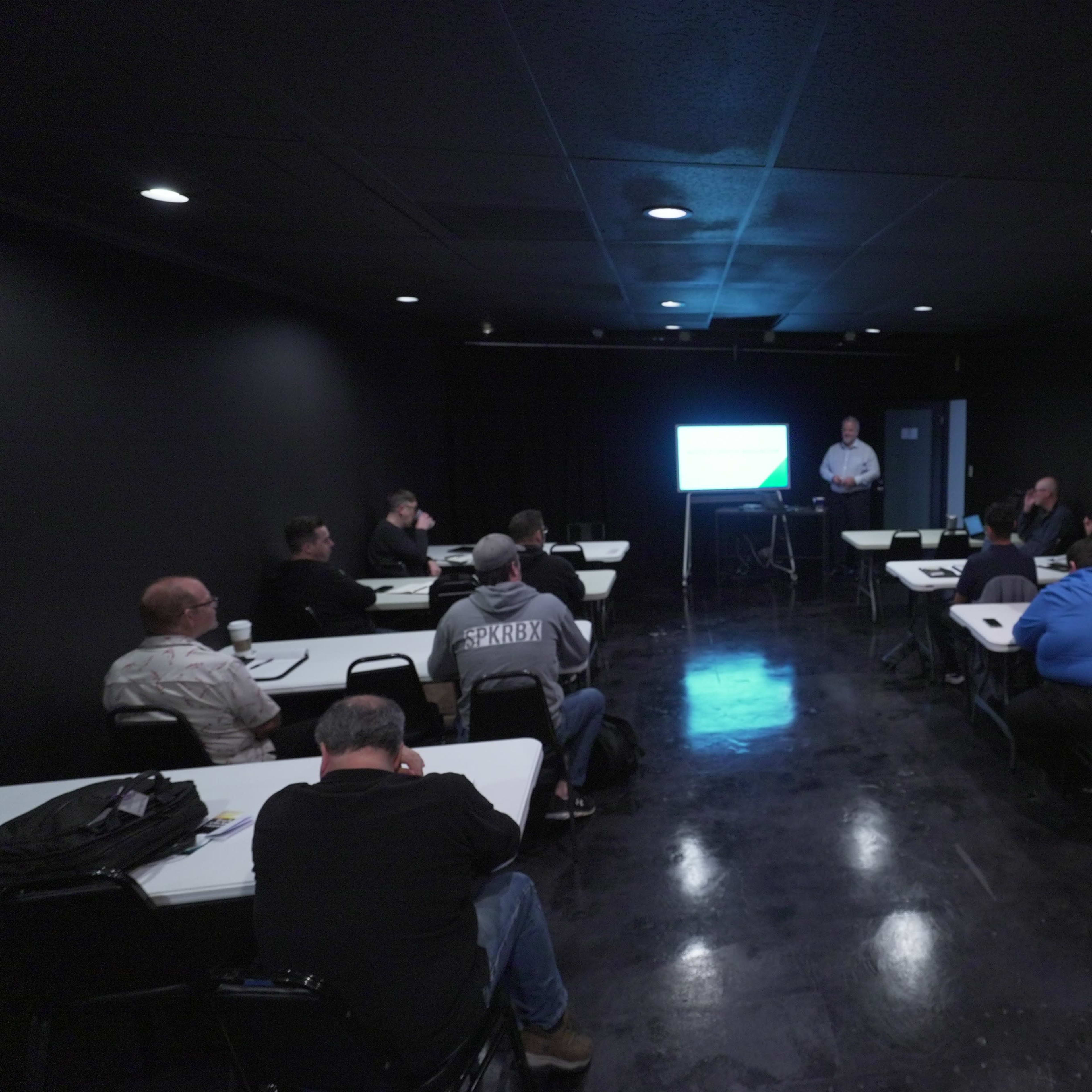 A group of people sits in a dimly lit conference room, attentively listening to a presenter near a screen displaying a presentation.