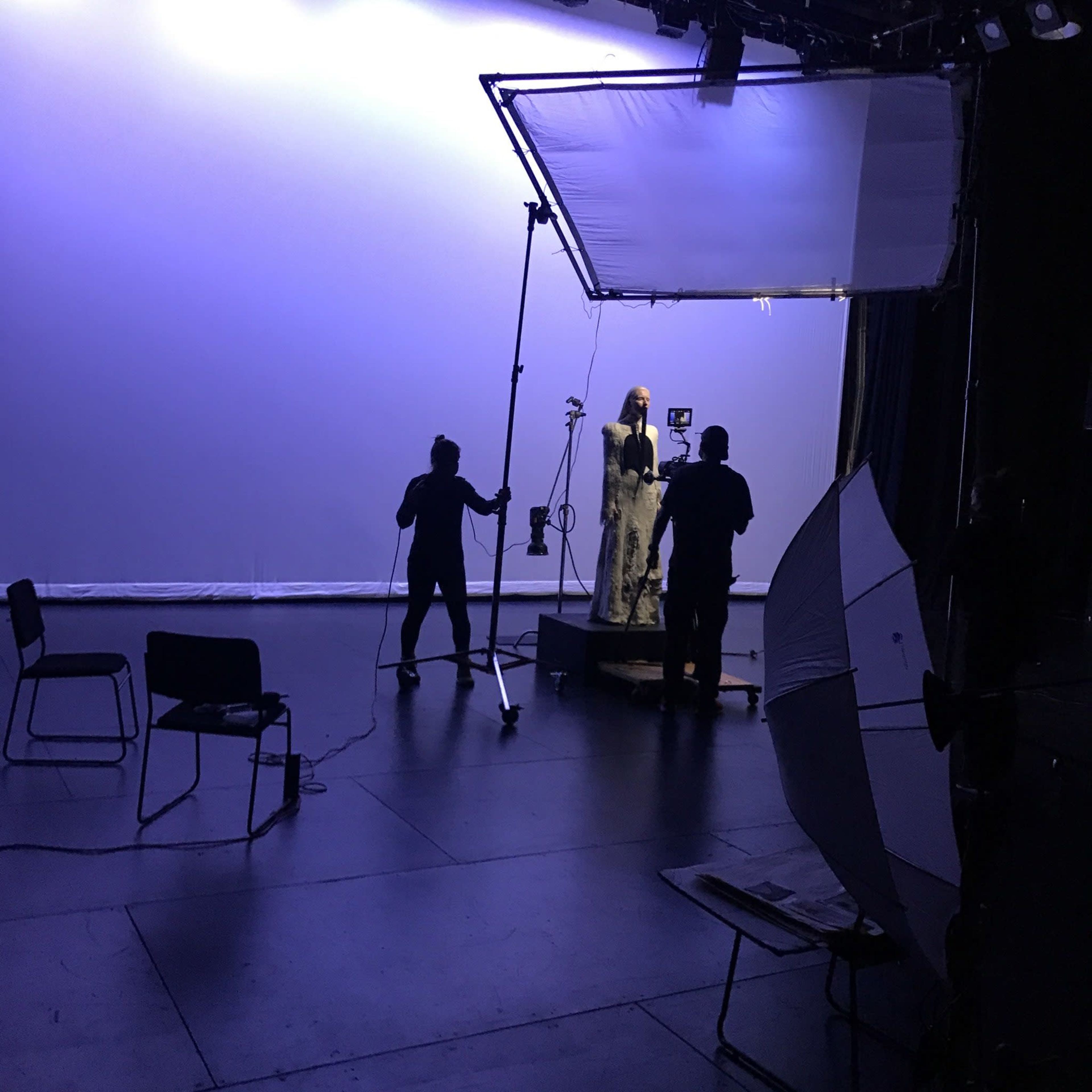 A film crew is setting up lighting and equipment in a studio for a shoot featuring a model.