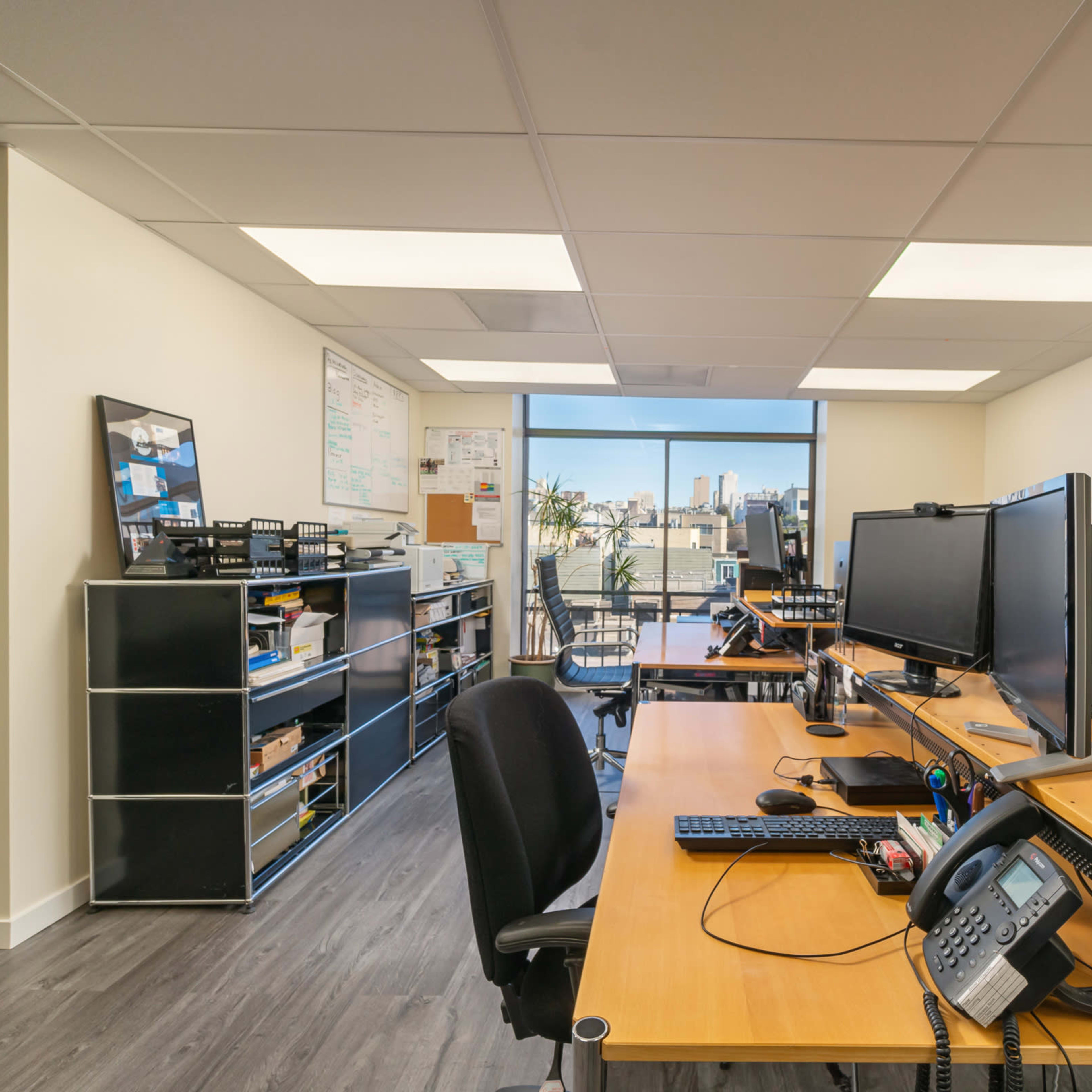A modern office space with two large computer monitors on a wooden desk, shelves filled with office supplies, and a view of the city skyline through a large window.