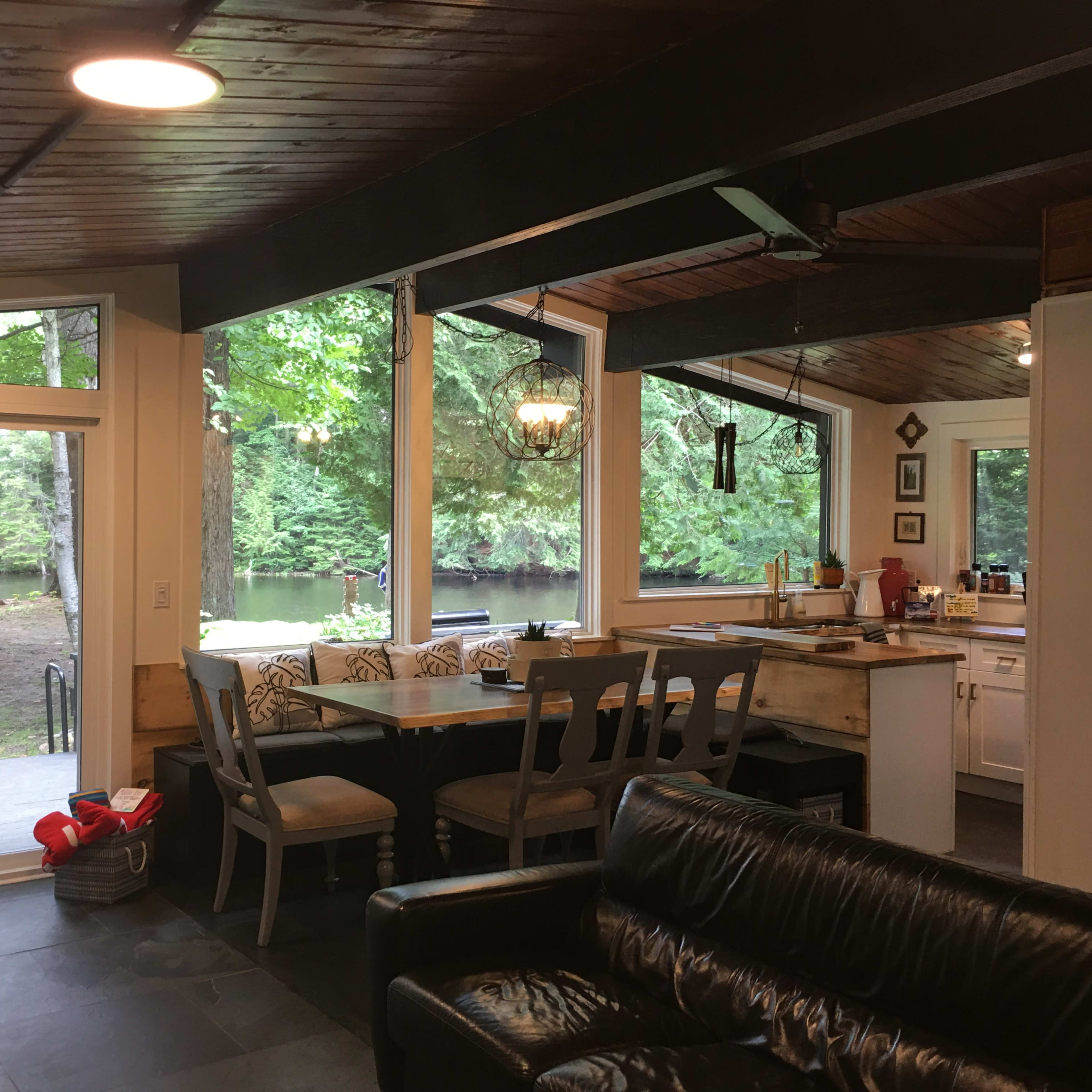 A bright, open kitchen area with a dining table, overlooking a lake and wooded landscape through large windows.