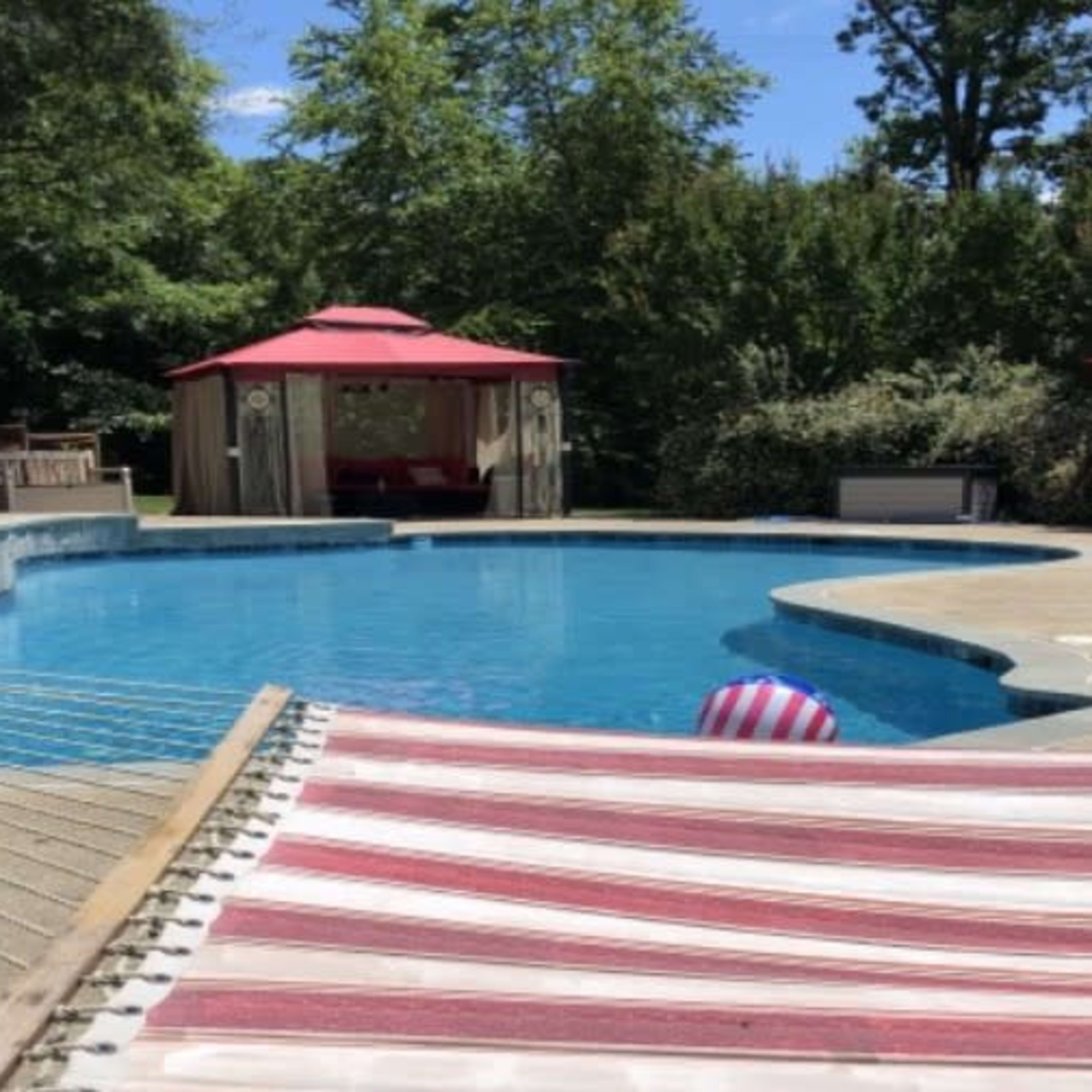 A backyard pool area with a blue swimming pool, two red canopies, lounge chairs, and a hammock in the foreground.