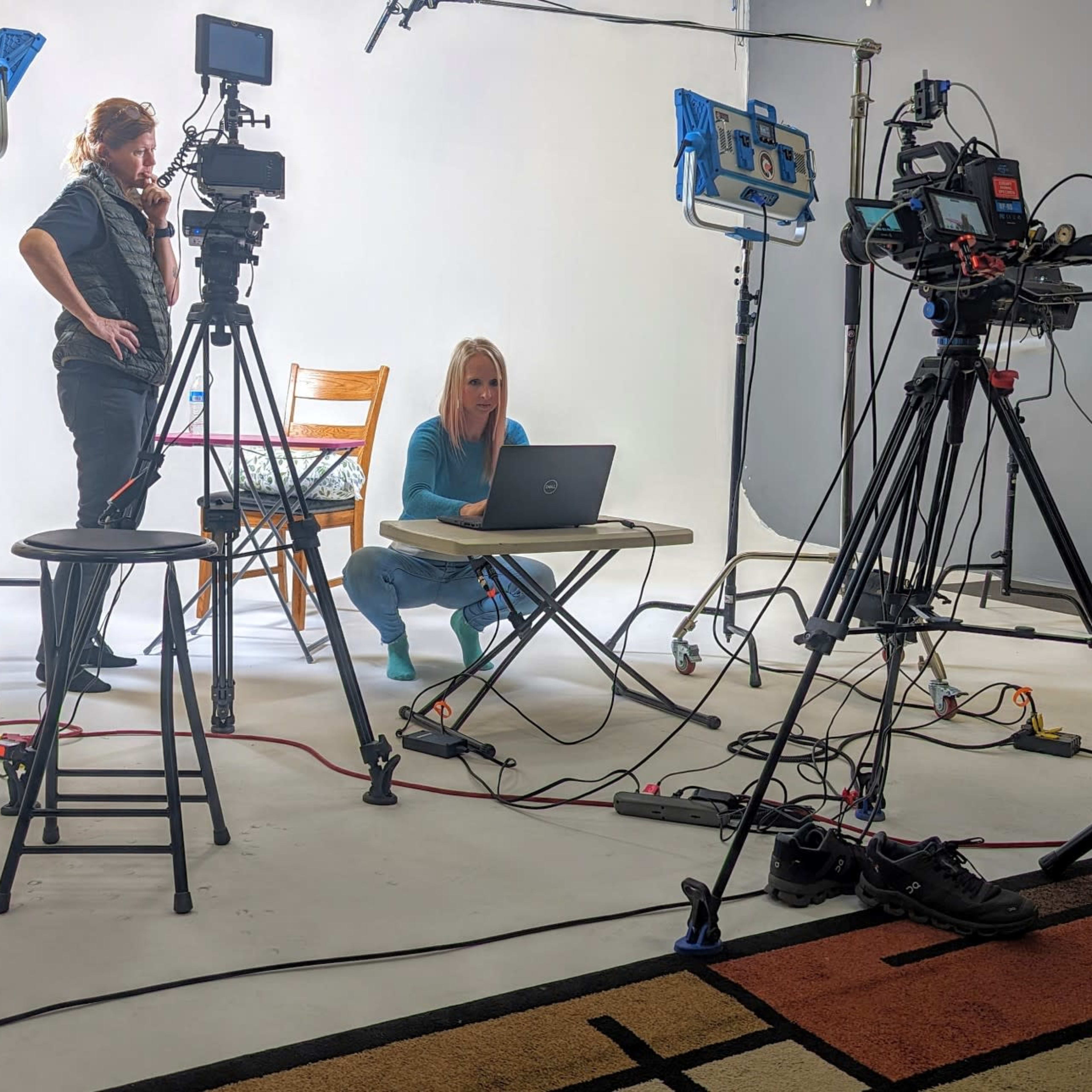 A woman sits at a table with a laptop while a man stands nearby, surrounded by multiple cameras and studio lights.
